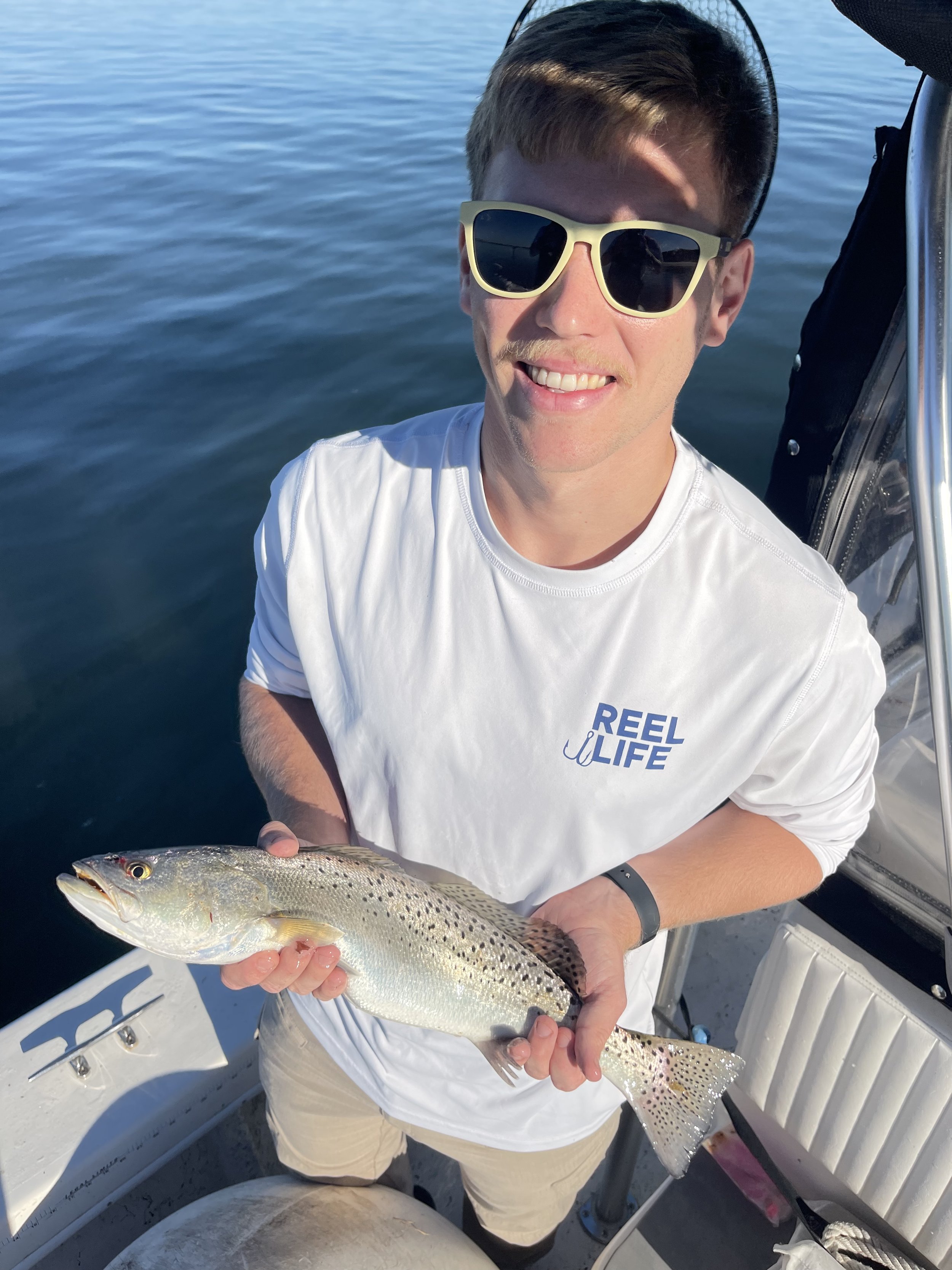 Young man wearing sunglasses and a white t-shirt holding a freshly caught fish on a boat over a calm body of water.