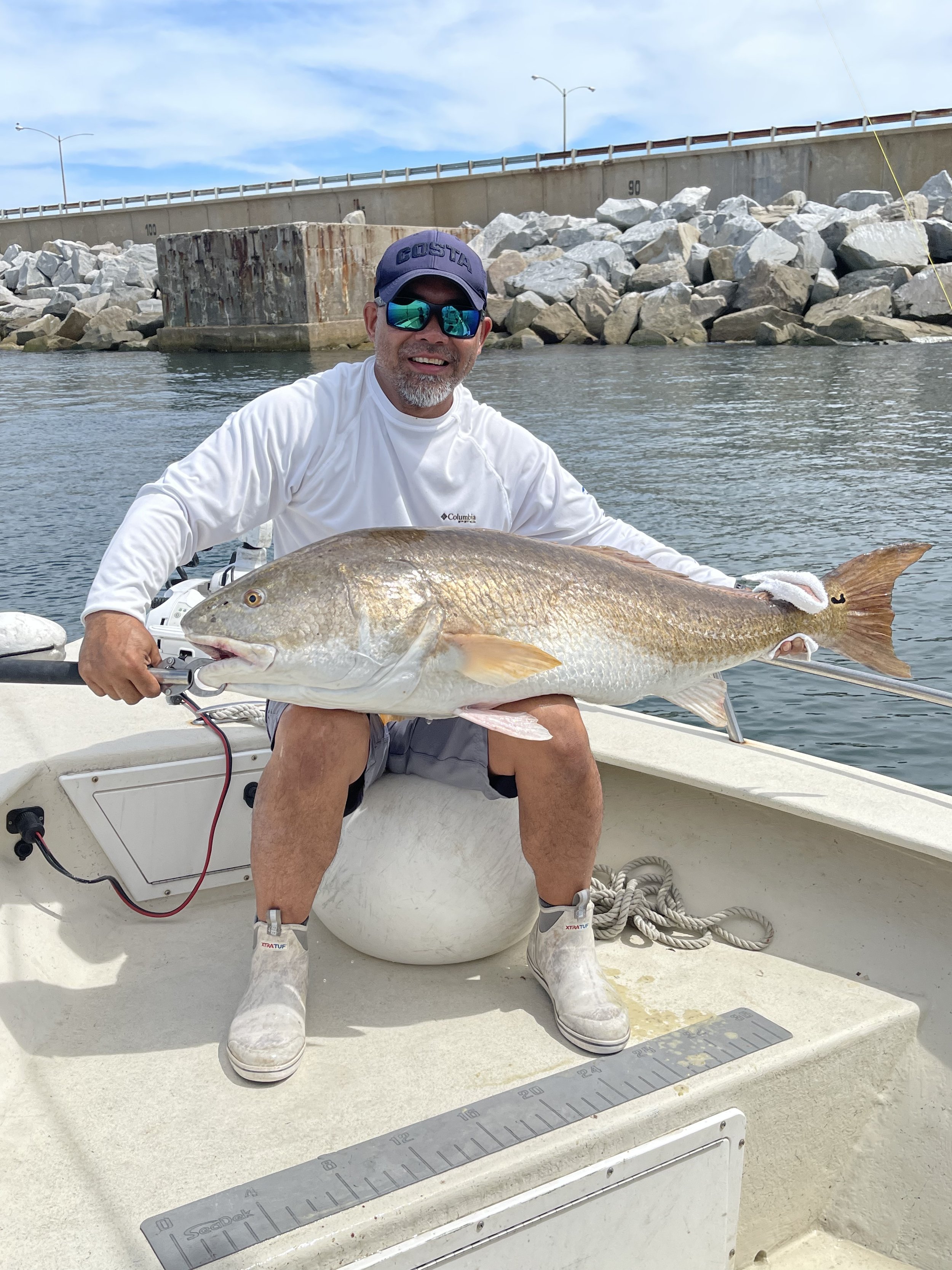 A man sitting on a boat holding a large fish he caught, with a stone and concrete structure and a bridge in the background.