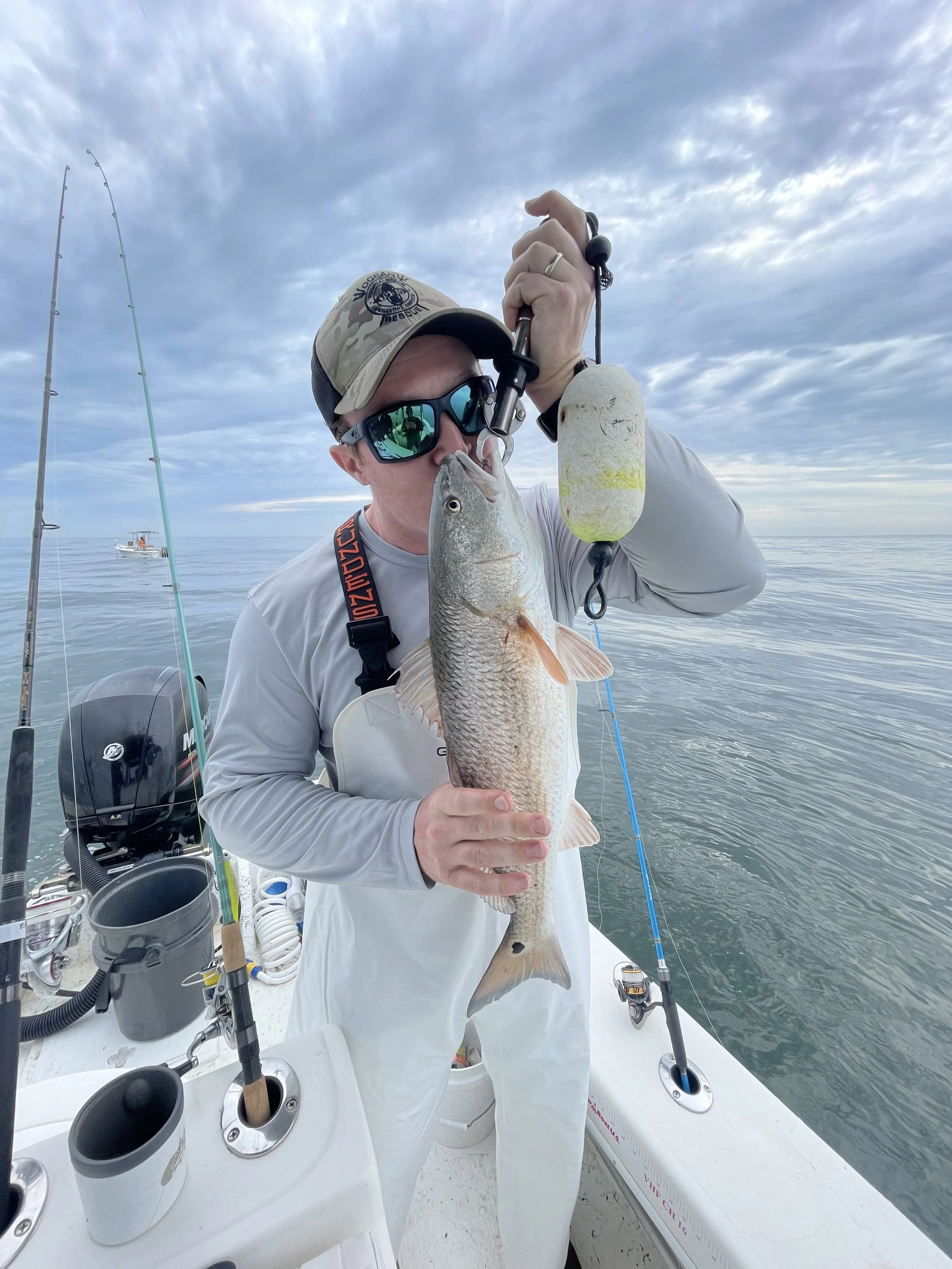 Man holding a large fish on a boat with fishing rods and ocean in the background.