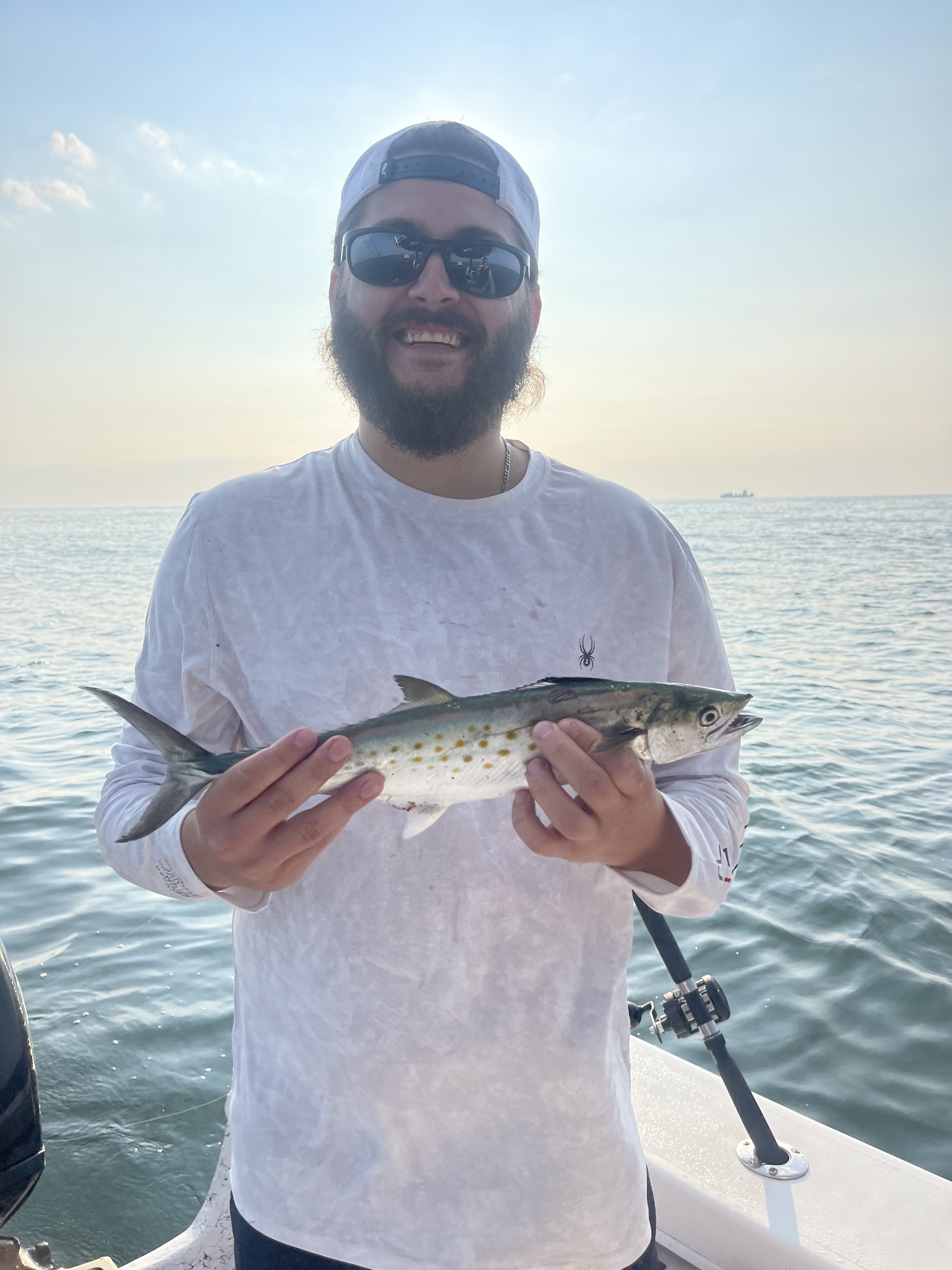 A man with a beard, sunglasses, and a backward baseball cap holding a fish on a boat, with the ocean and distant ship in the background.