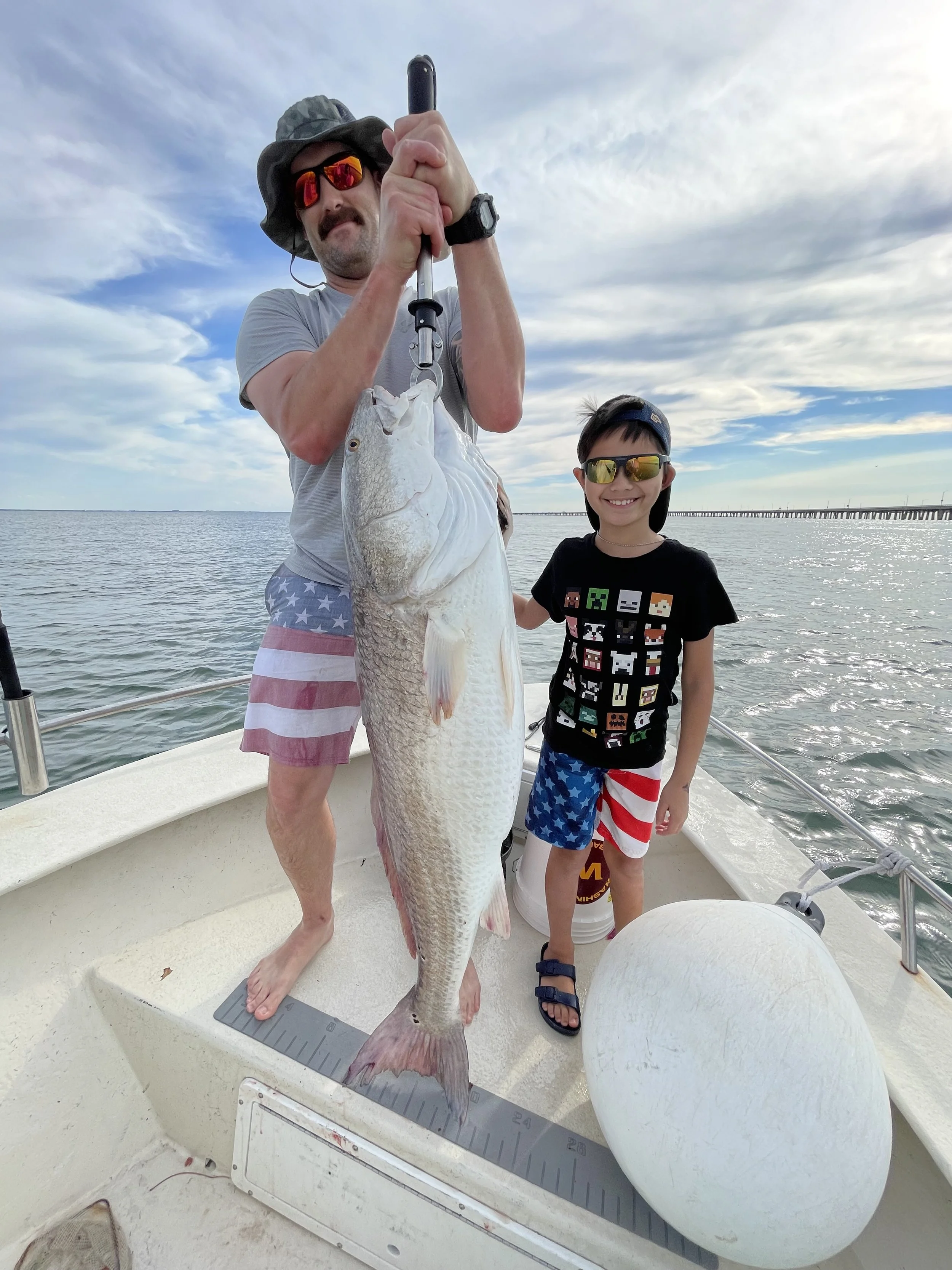 A man and a boy on a boat holding a large fish. The man is holding the fish with a gripping tool, and both are smiling. The man is wearing sunglasses, a hat, and shorts with an American flag pattern. The boy is wearing sunglasses, a t-shirt with colo