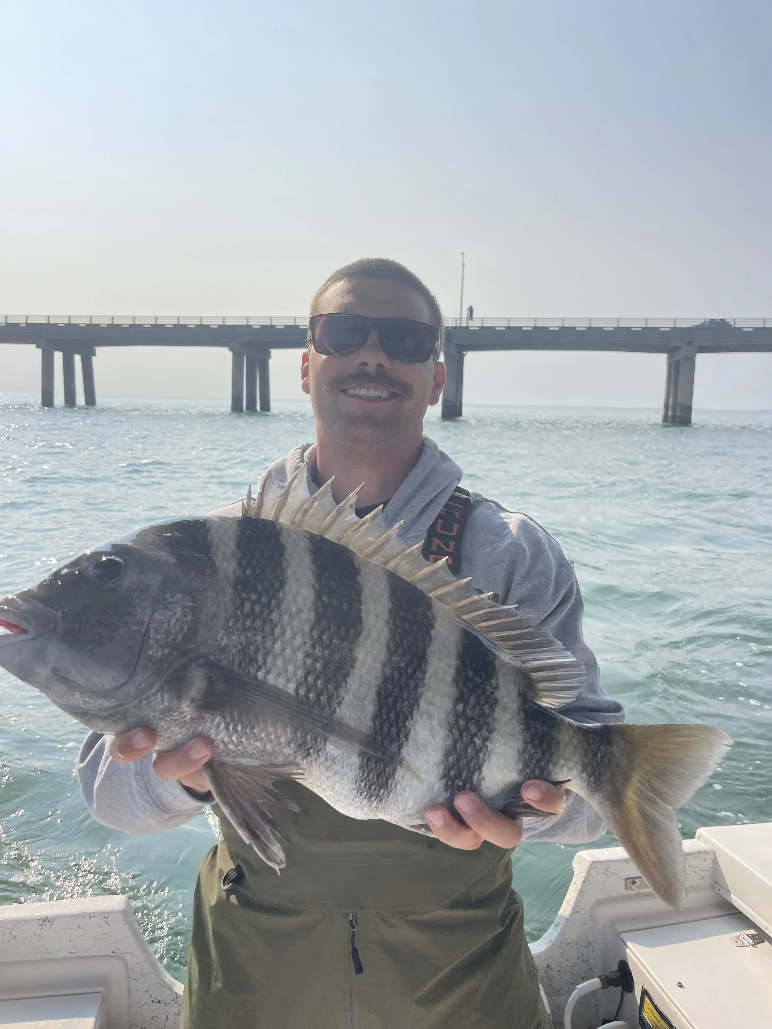 A man in sunglasses smiling while holding a large fish on a boat with a pier in the background.
