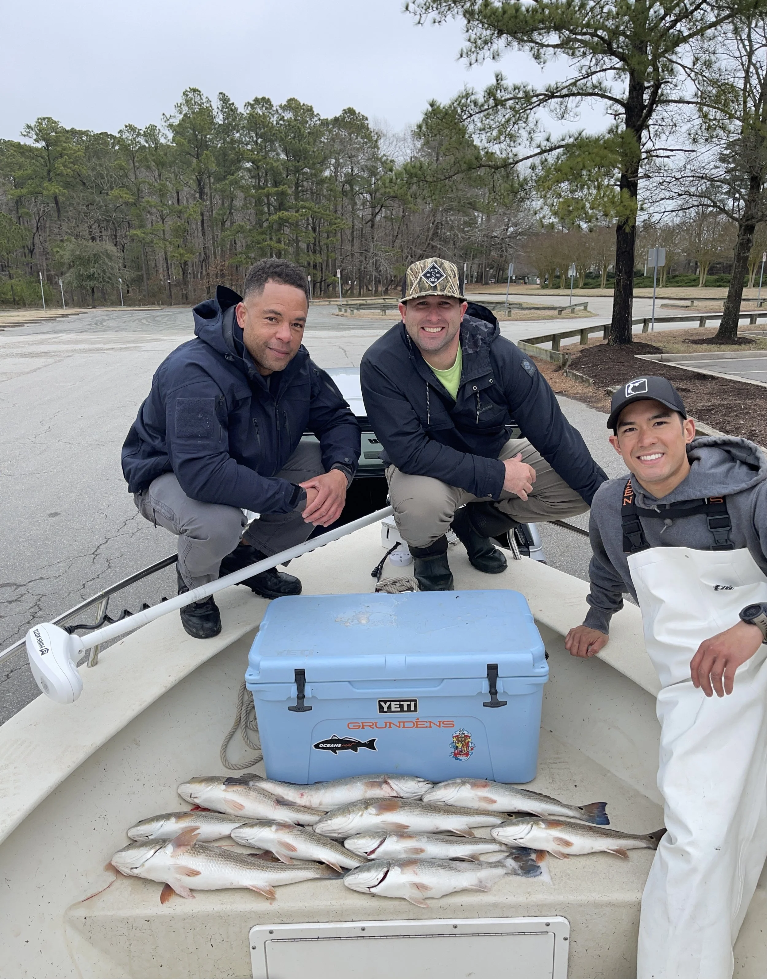 Three men on a boat with caught fish and fishing gear, smiling for the camera, in an outdoor parking lot with trees in the background.