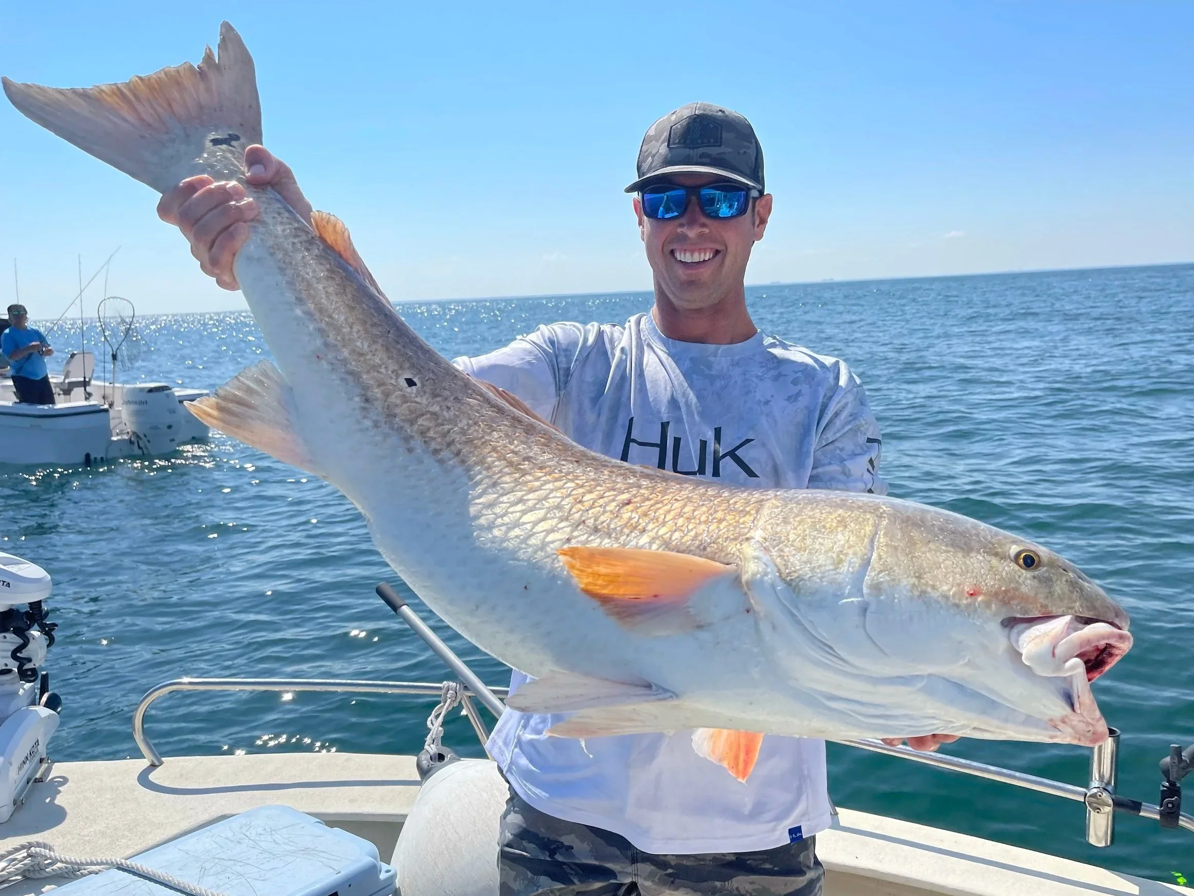 A smiling man wearing sunglasses and a cap holding a large fish on a boat in the ocean, with another person fishing in the background.