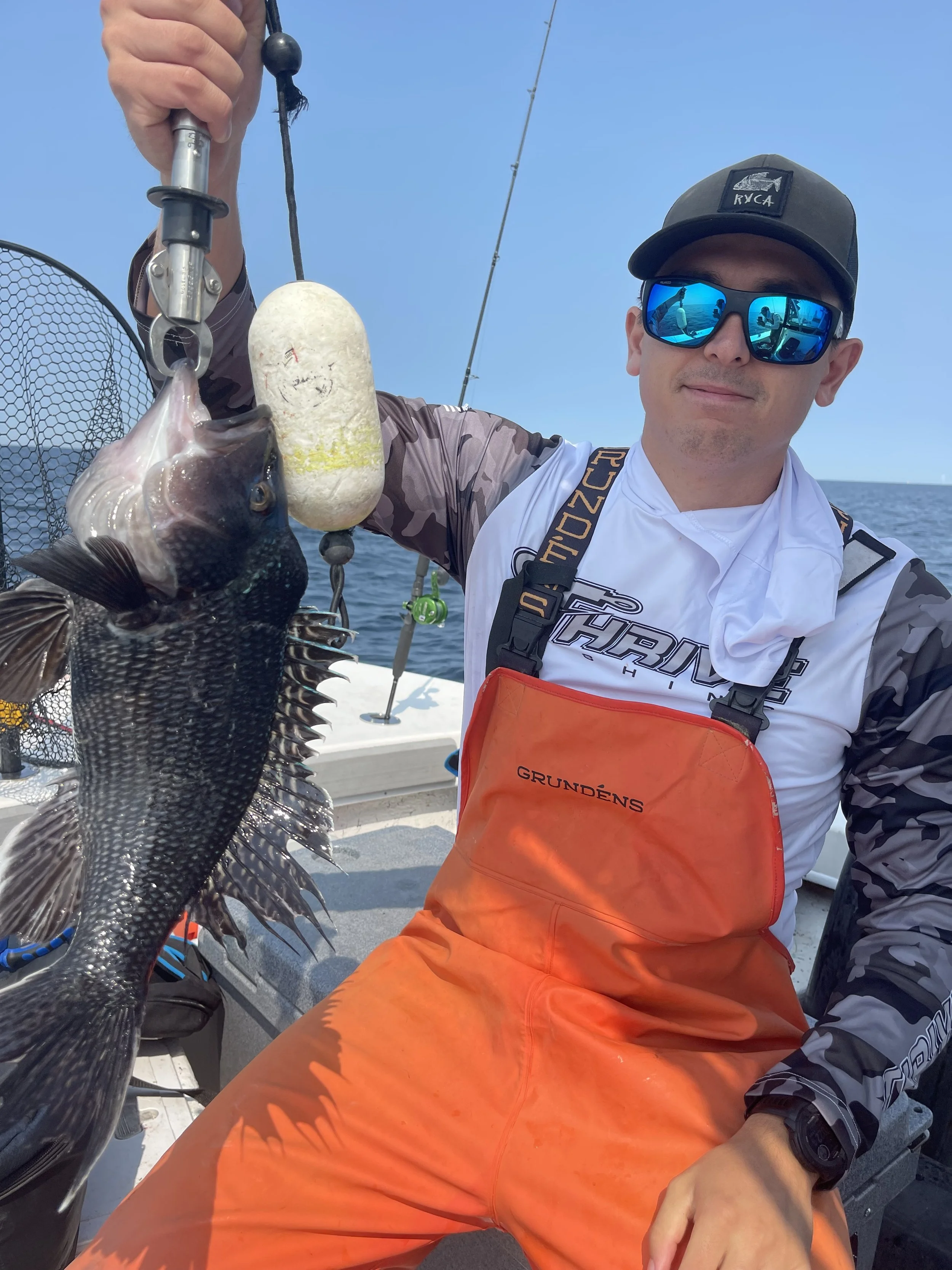 Man on a boat holding a caught fish, wearing reflective sunglasses, a black cap, an orange bib with 'GRUNDÉNS' written on it, and a camouflage shirt.