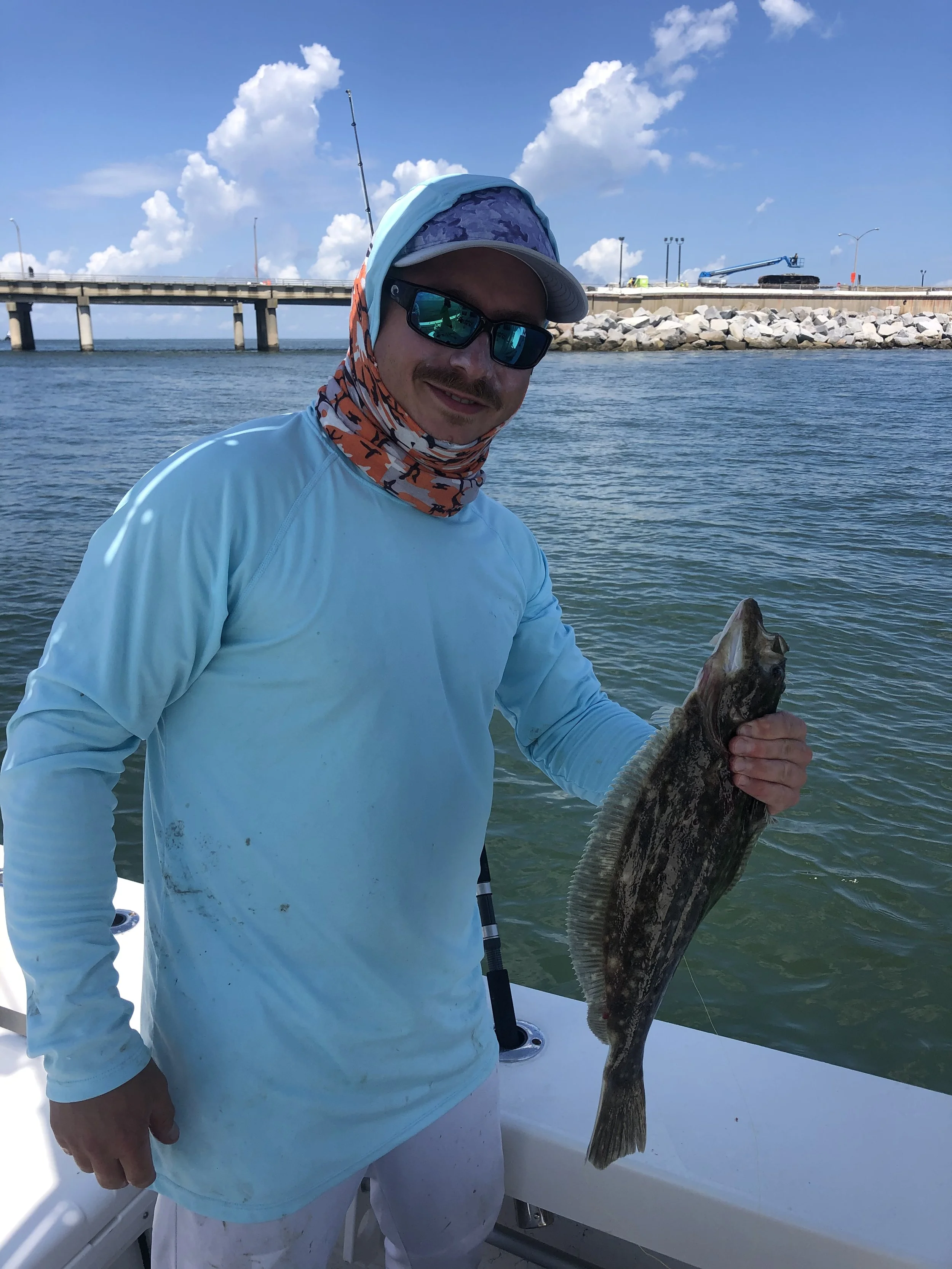 A man wearing sunglasses, a light blue long-sleeve shirt, and a floral neck gaiter holds a large fish on a boat. The background shows a bridge, rocks, and blue sky with white clouds.
