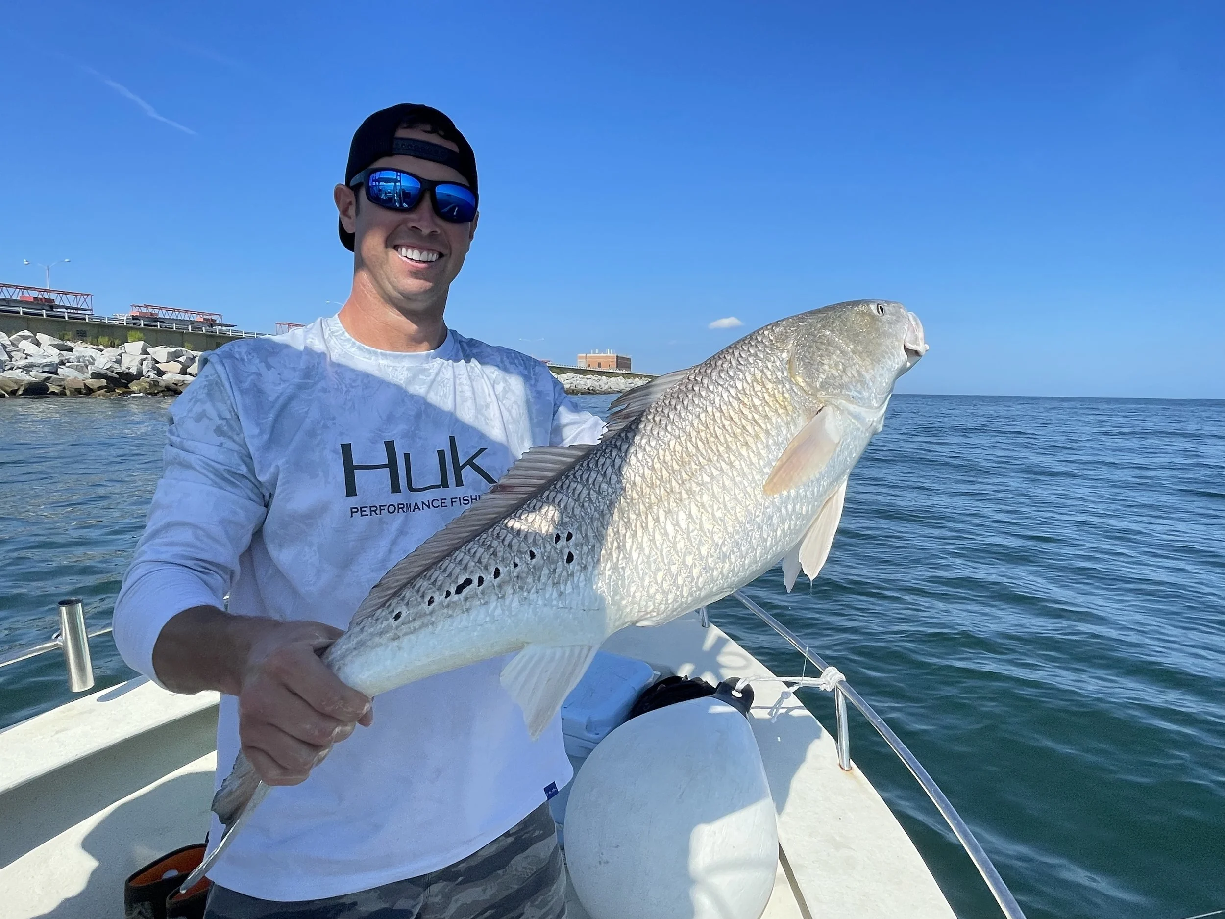 Smiling man wearing sunglasses and a white Huk Performance Fishing shirt holding a large fish on a boat in the water with a clear blue sky in the background.