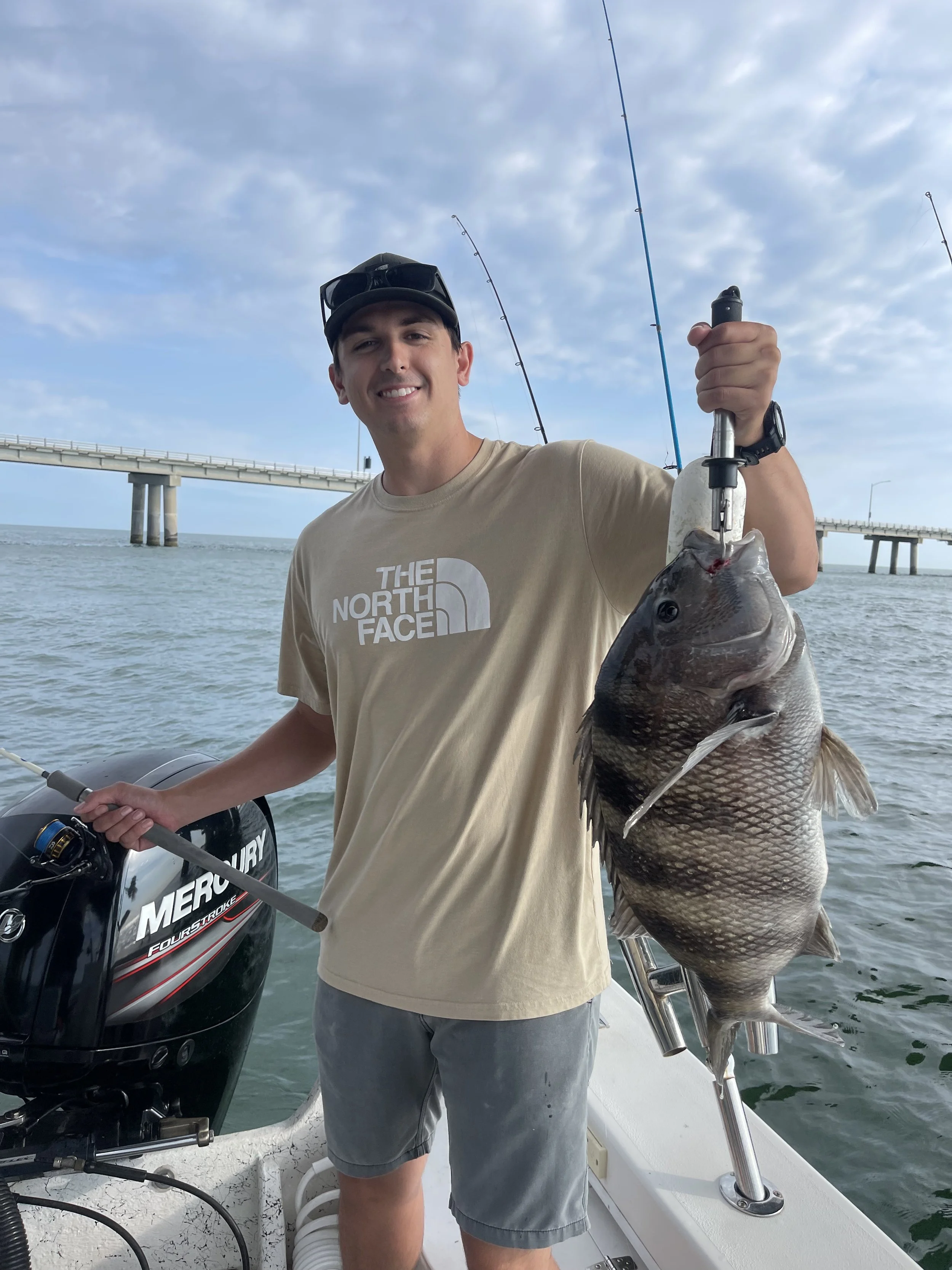 A man smiling on a boat holding a large fish he caught, with fishing poles in the background and an ocean pier.