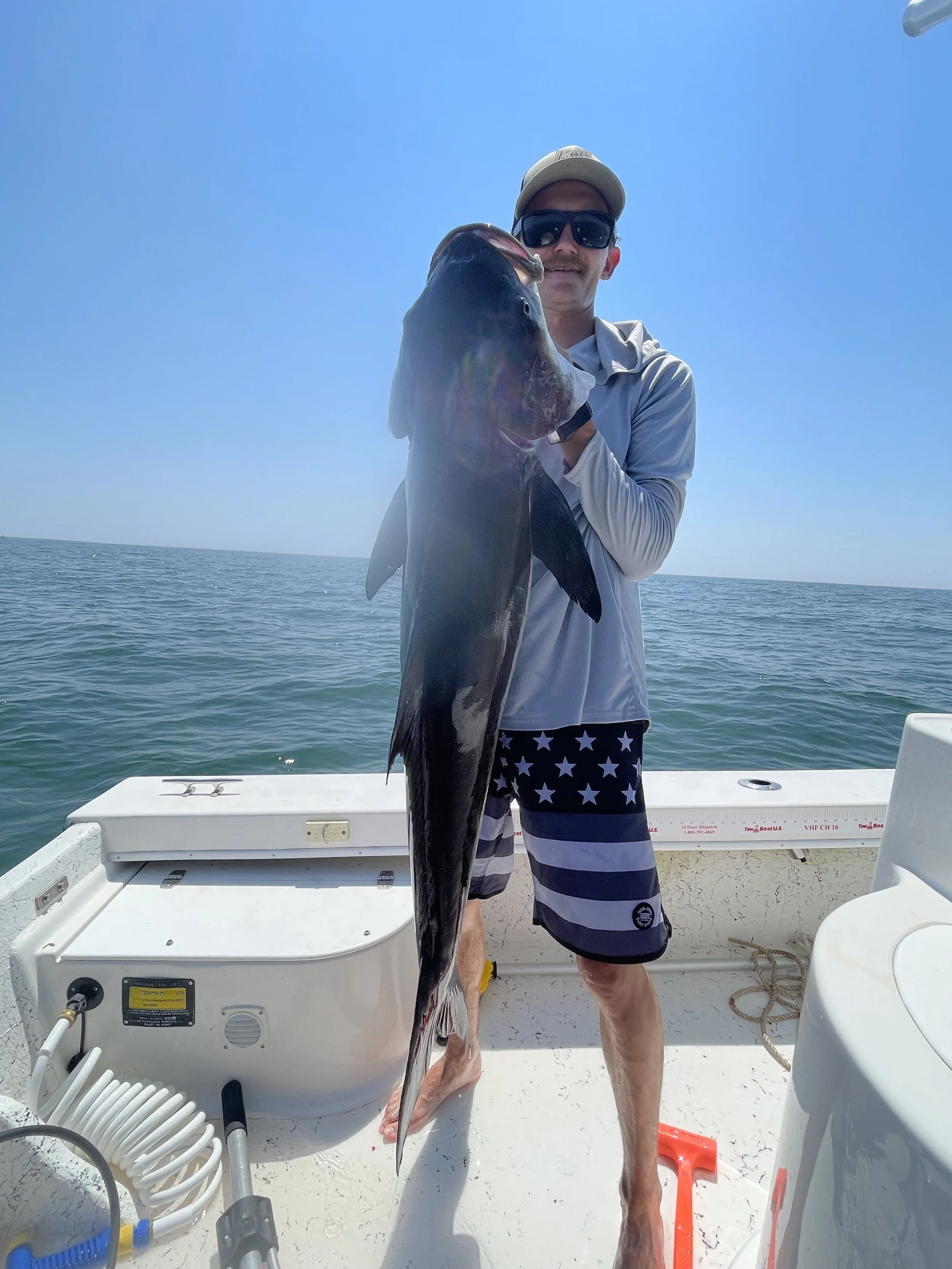 Man holding a large fish on a boat in the ocean on a sunny day, wearing sunglasses, a cap, a light jacket, and star-spangled shorts.