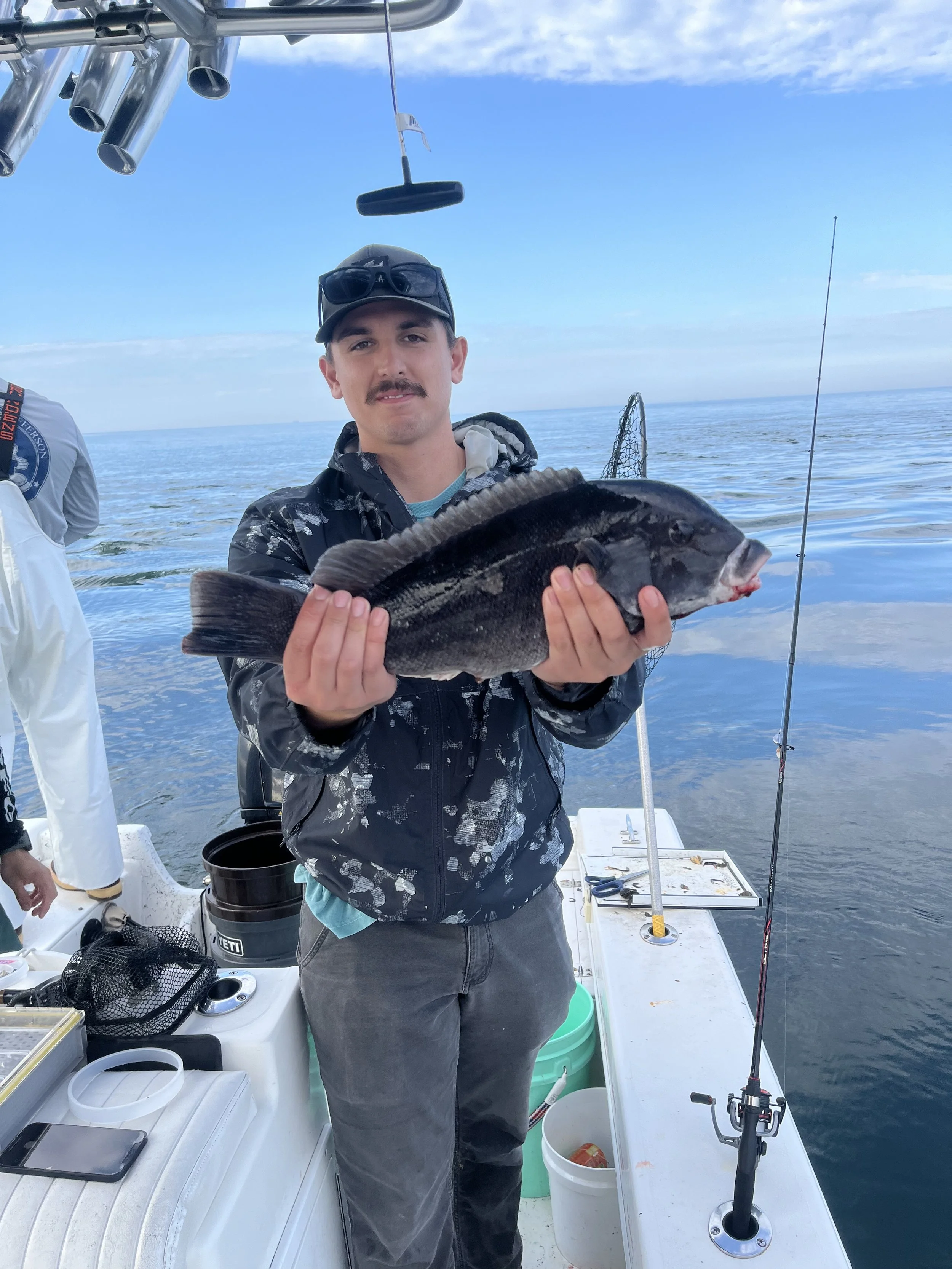 A man with a mustache wearing sunglasses, a hat, and a black jacket holds a large fish on a boat. The boat is on open water with a fishing rod and various fishing gear visible nearby.