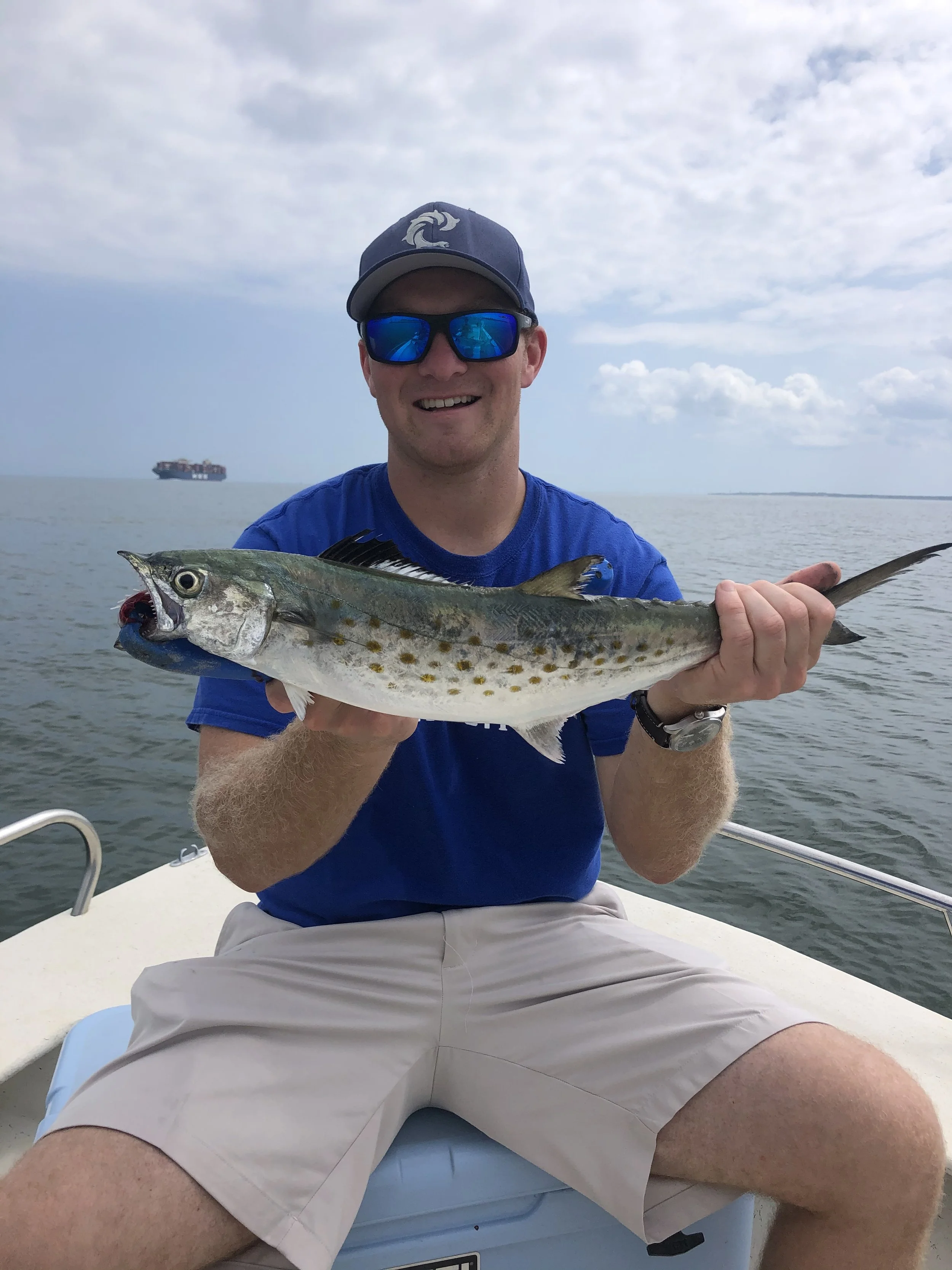 Man in sunglasses and a blue shirt holding a fish on a boat with water and sky in the background.