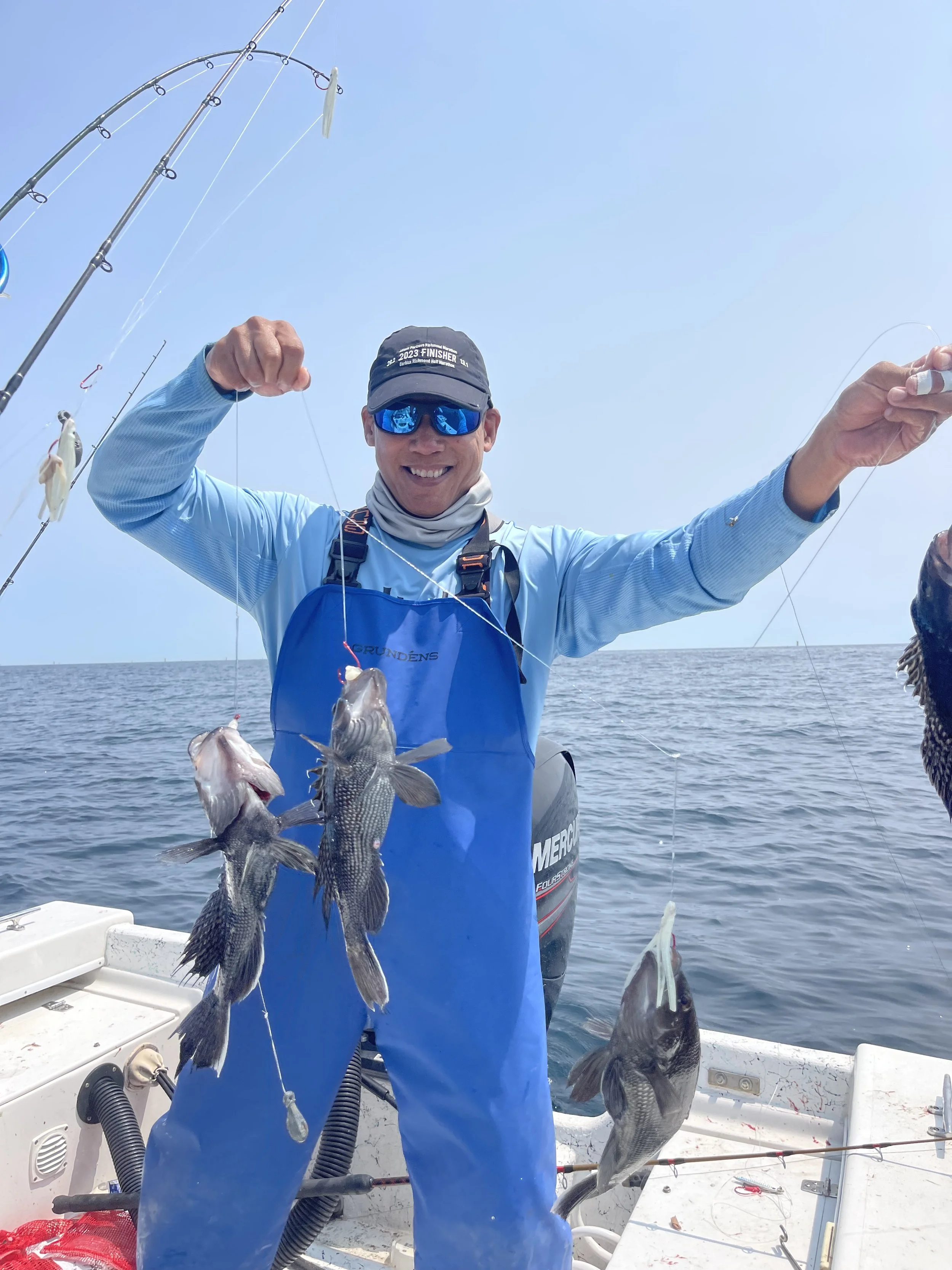 Smiling man posing on a boat holding a string with three caught fish, wearing sunglasses and a blue outfit with a cap and neck gaiter, with other fishing rods visible against a calm ocean background.