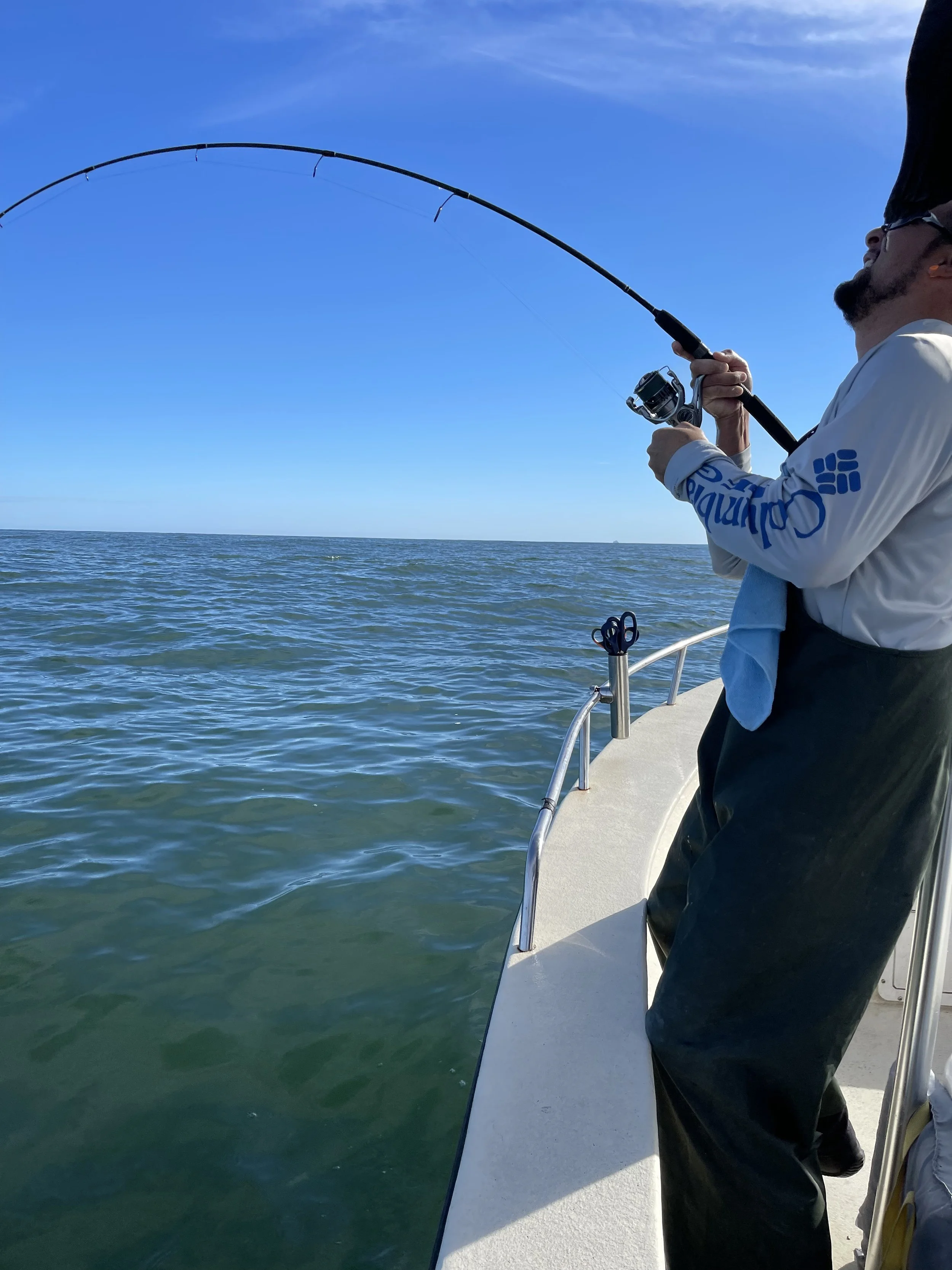 Man fishing on a boat in the ocean, casting his fishing line into the water with clear blue sky overhead.