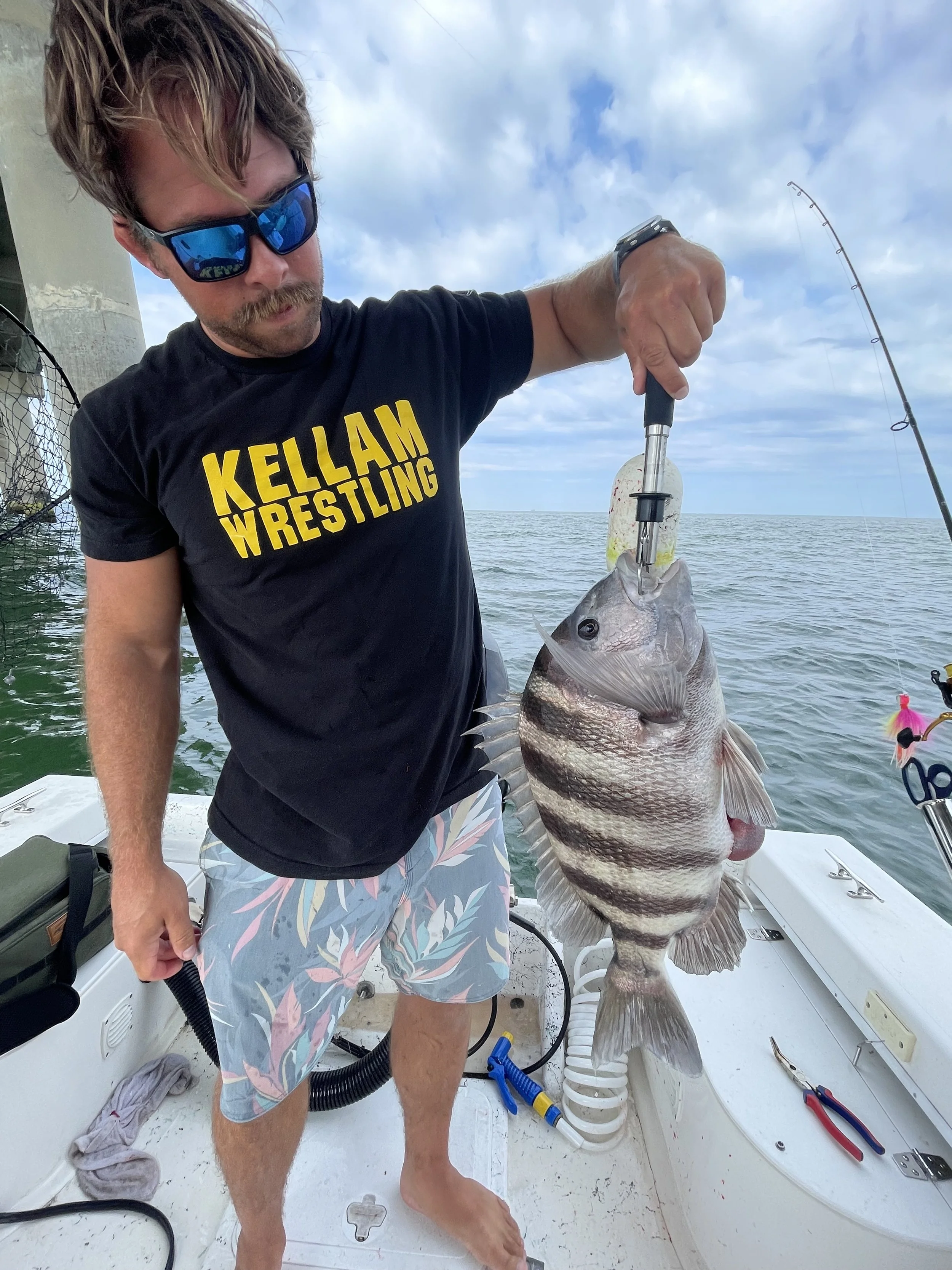 Man wearing sunglasses, a black T-shirt with yellow text and patterned shorts, holding a large fish on a boat with fishing gear and the ocean in the background.