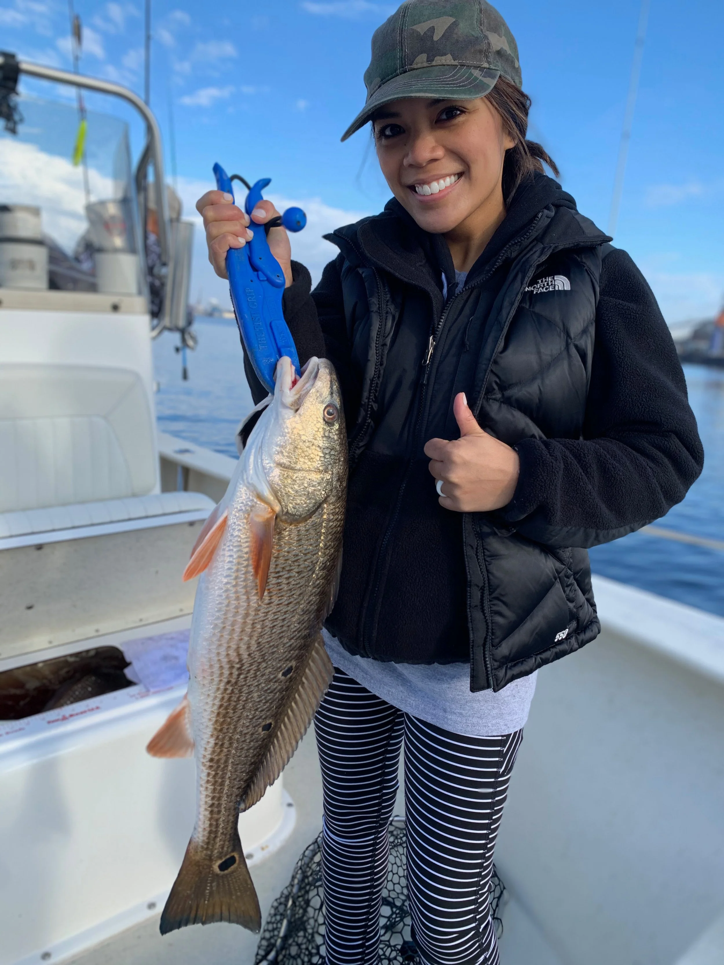 Woman on a boat holding a large fish with a blue gripping tool, smiling and giving a thumbs up, with water and a clear sky in the background.
