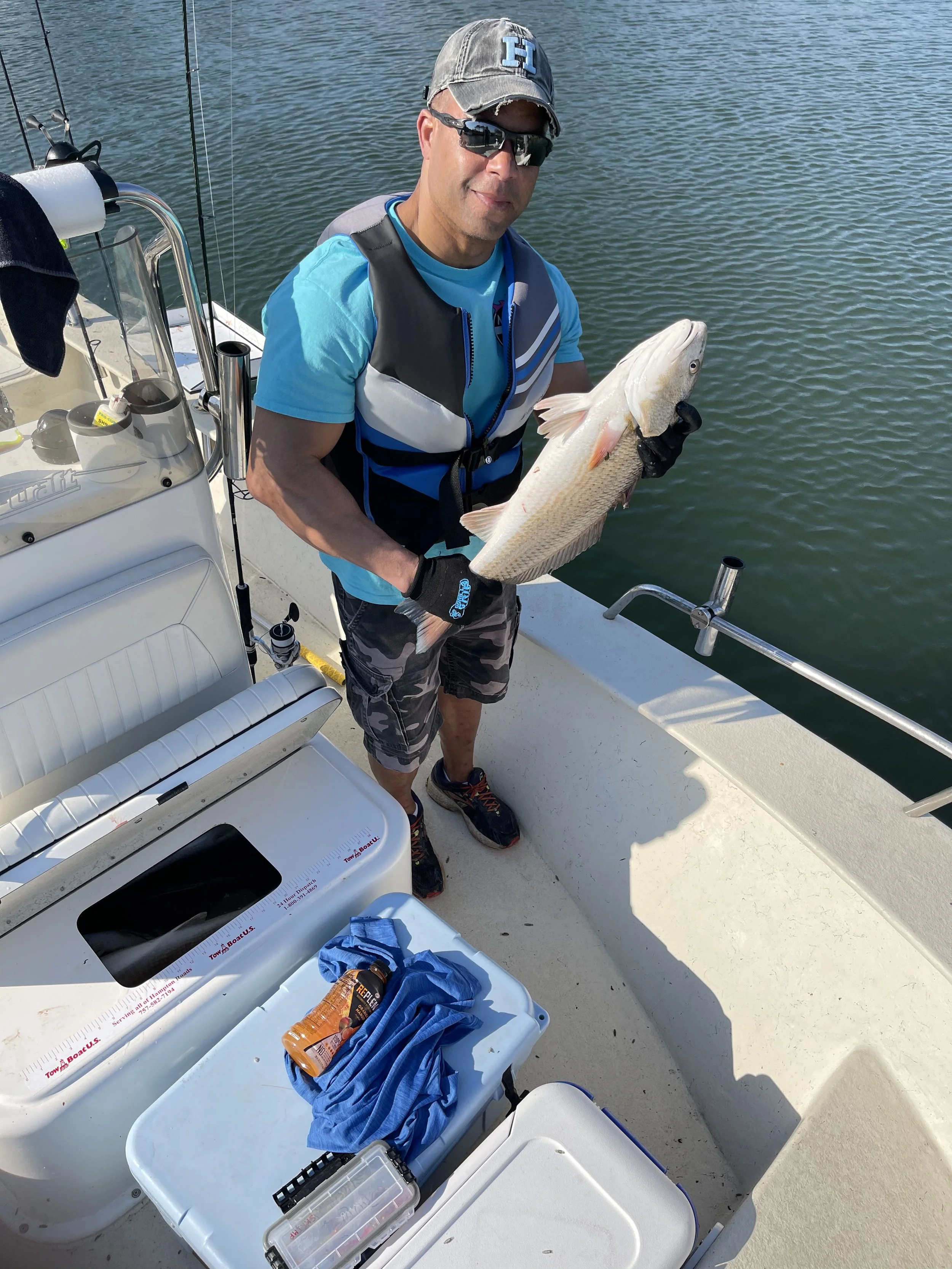 Man on a boat holding a large fish he caught, wearing sunglasses, a gray cap, a blue shirt, and a life vest.