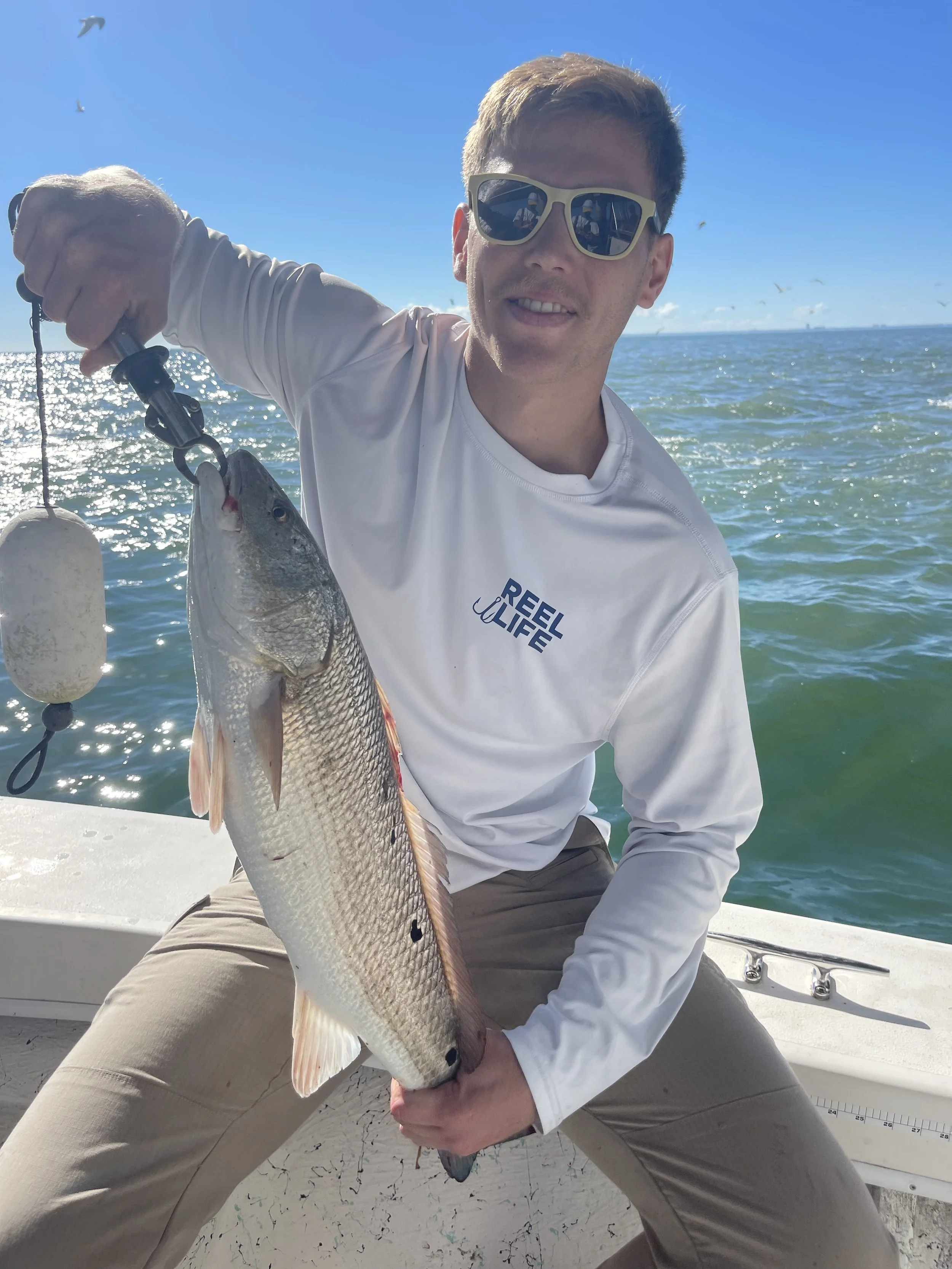 A man wearing sunglasses and a white long-sleeve shirt that says 'Reel Life' is sitting on a boat and holding a large fish he caught, with the ocean and a clear blue sky in the background.