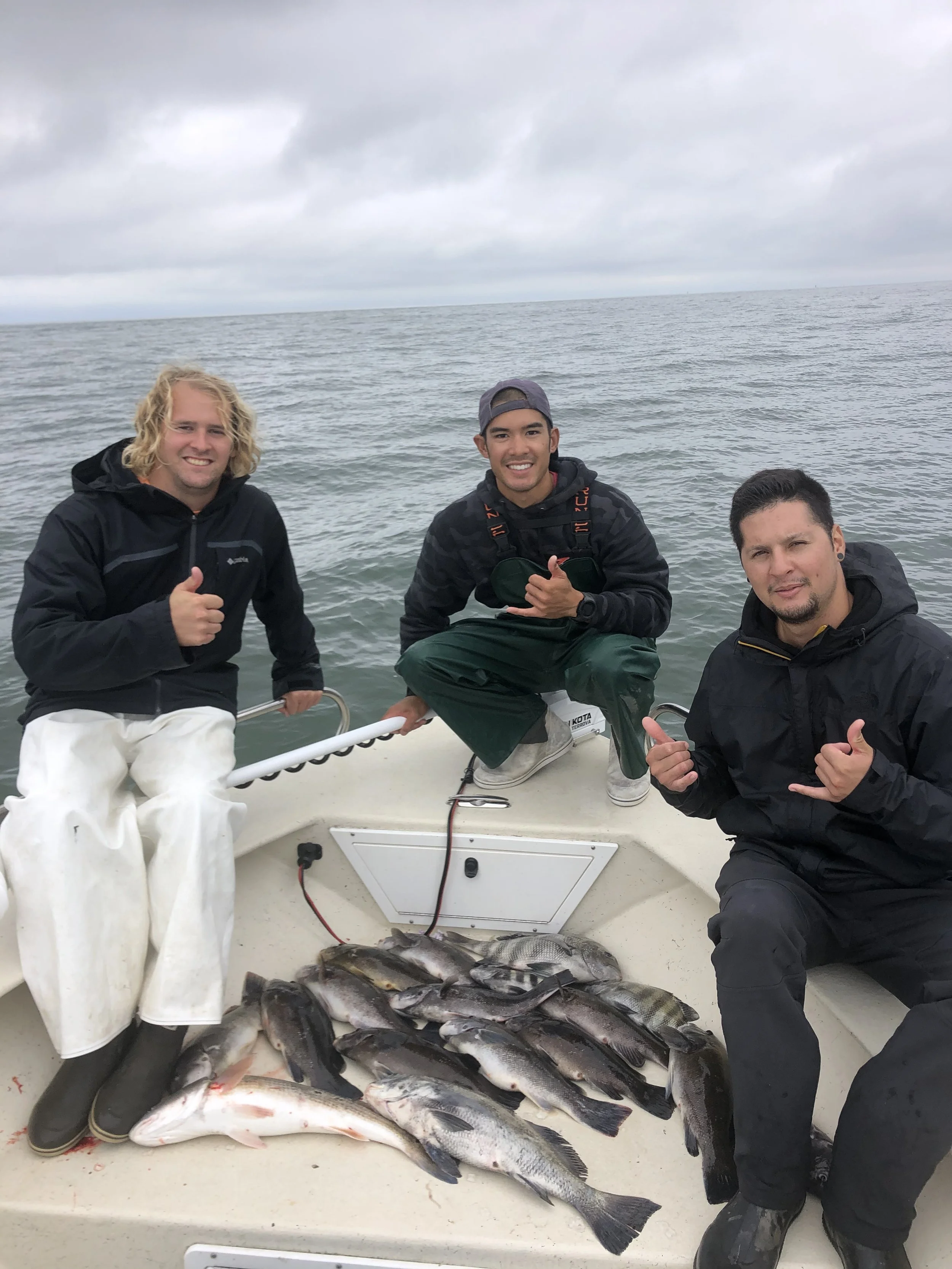 Three men on a boat with a catch of fish, giving thumbs up and smiling, on the open water with overcast skies.