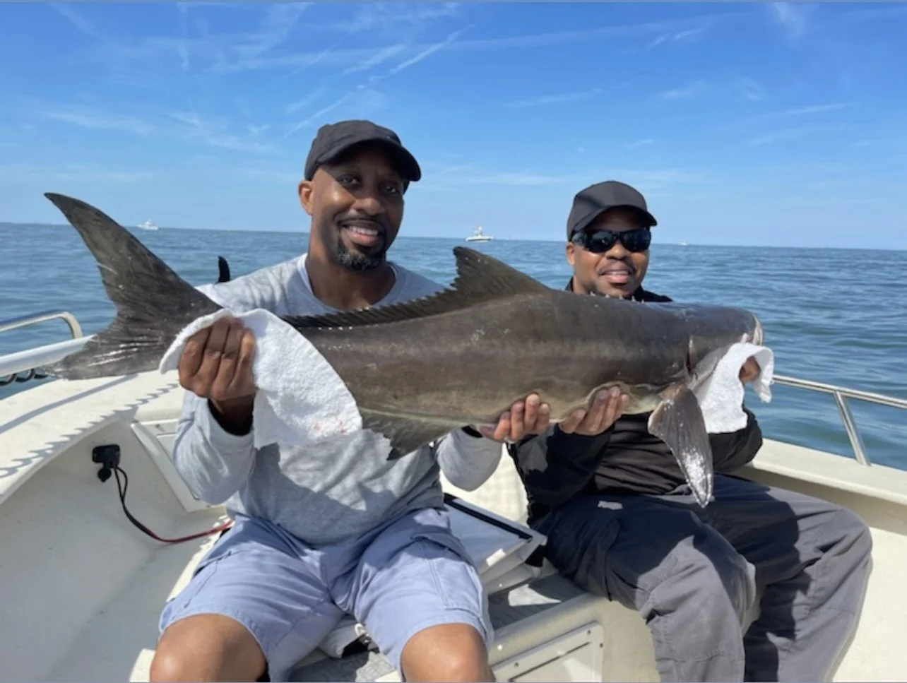 Two men sitting on a boat holding a large fish between them, with an ocean and clear blue sky in the background.
