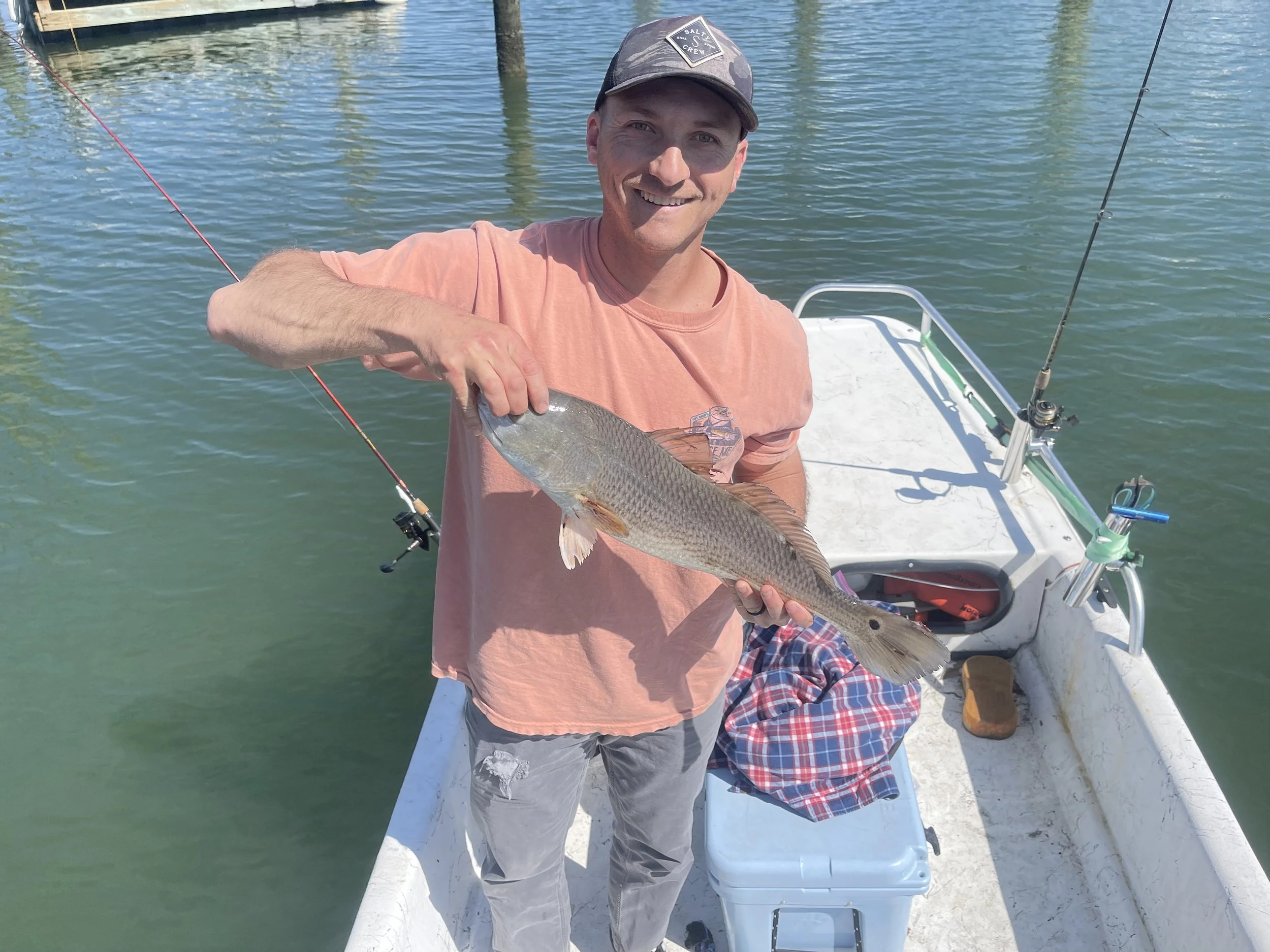 Man smiling on a boat holding a fish he caught, with fishing rods and water visible in the background.