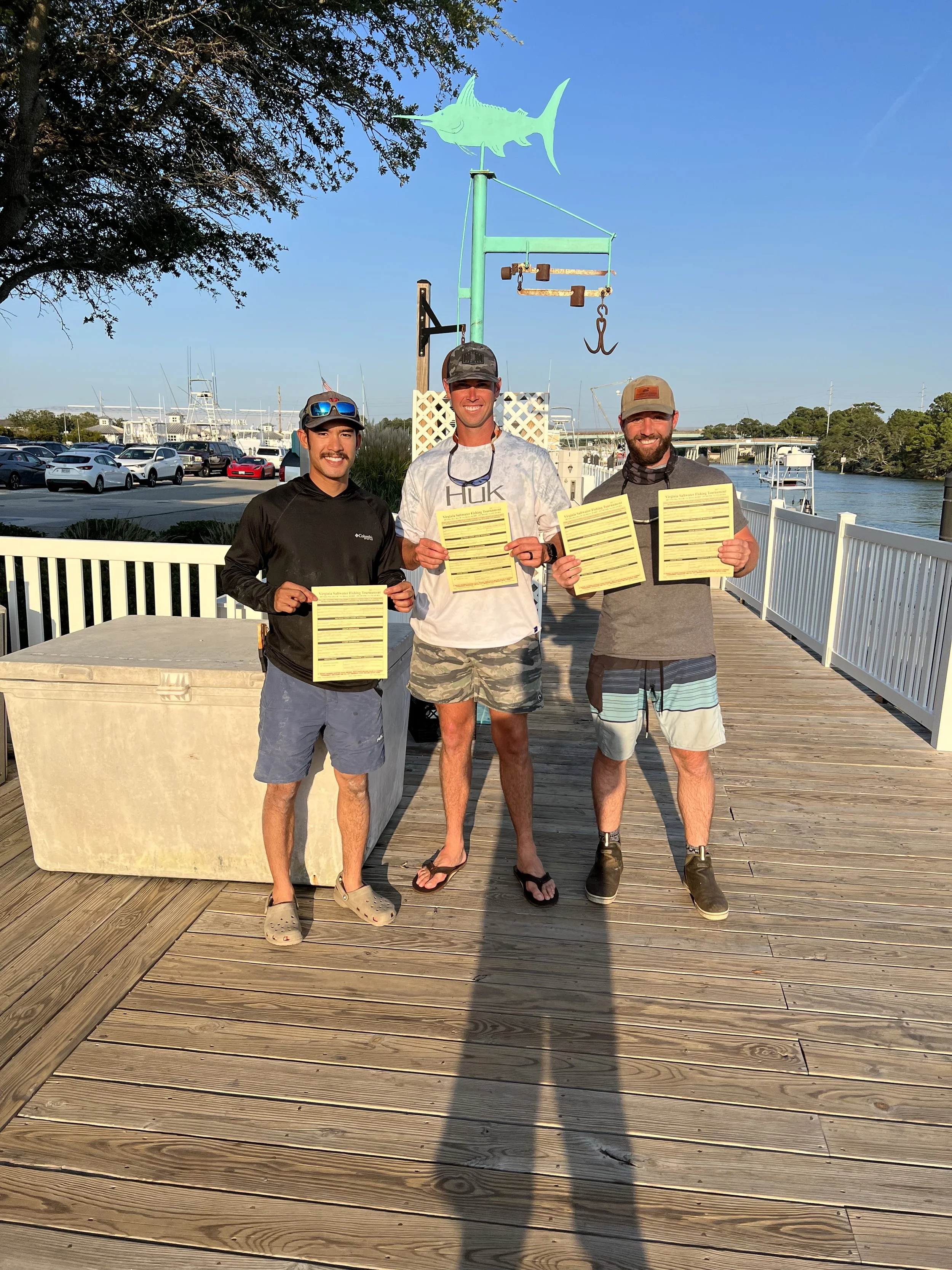 Three men standing on a wooden dock, holding fishing permits, with a decorative fish weather vane above them and a marina in the background.