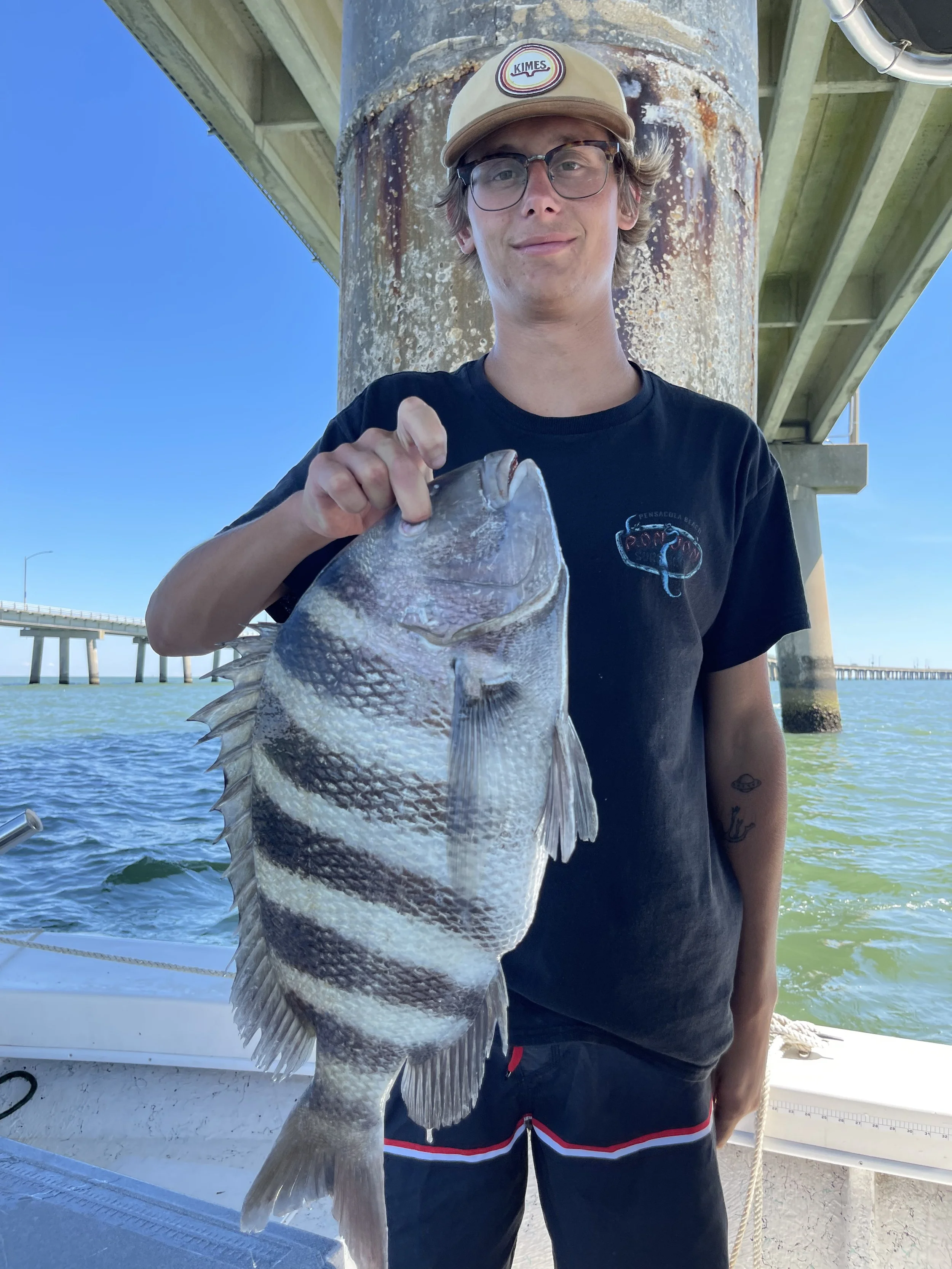 A young man wearing glasses, a beige cap with a logo, and a black t-shirt showing a fish graphic, standing on a boat under a bridge. He is holding a large fish with striped black and white markings, with the water and bridge in the background.