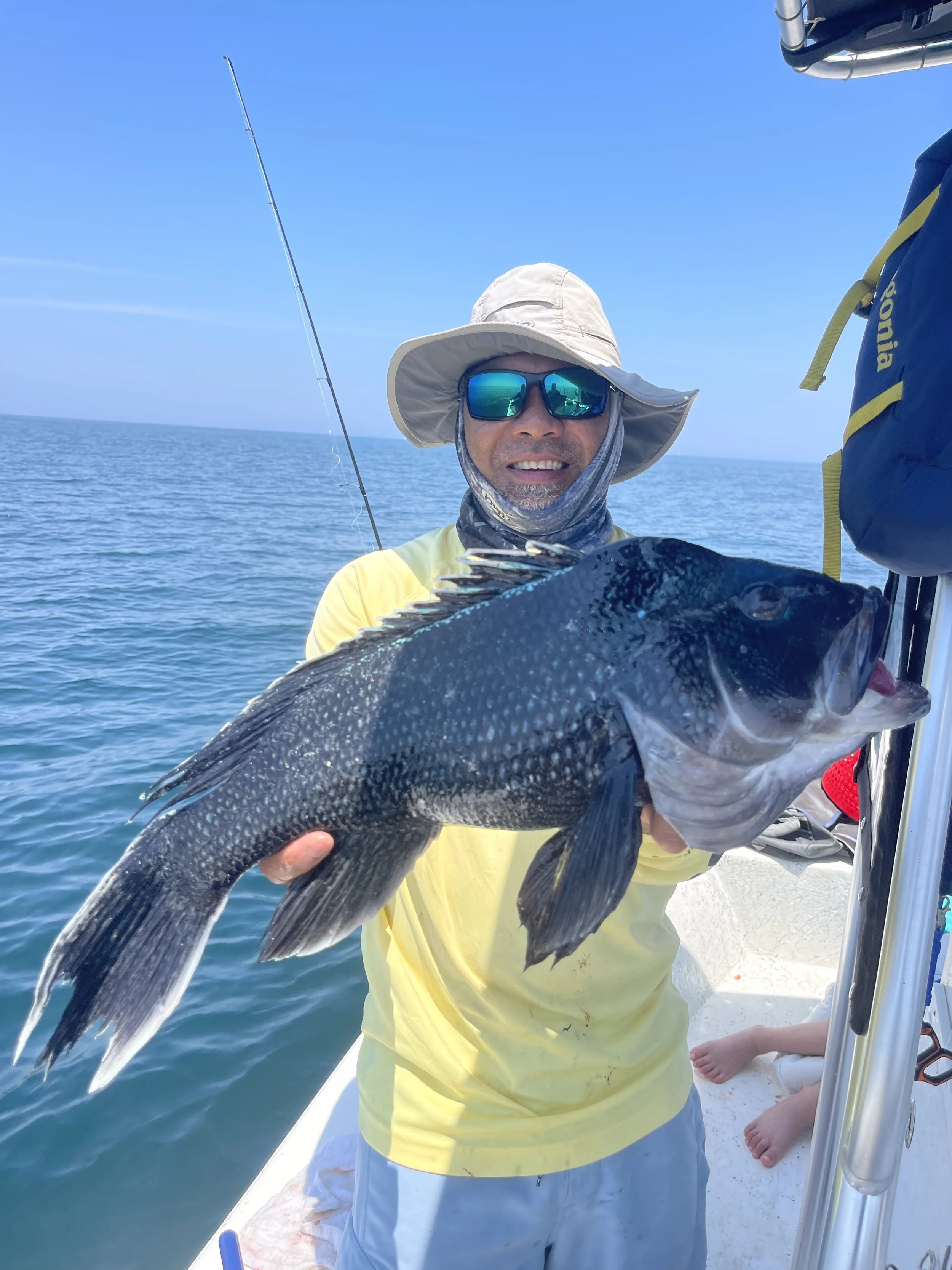 Man on a boat holding a large black fish he caught, surrounded by ocean and clear blue sky.