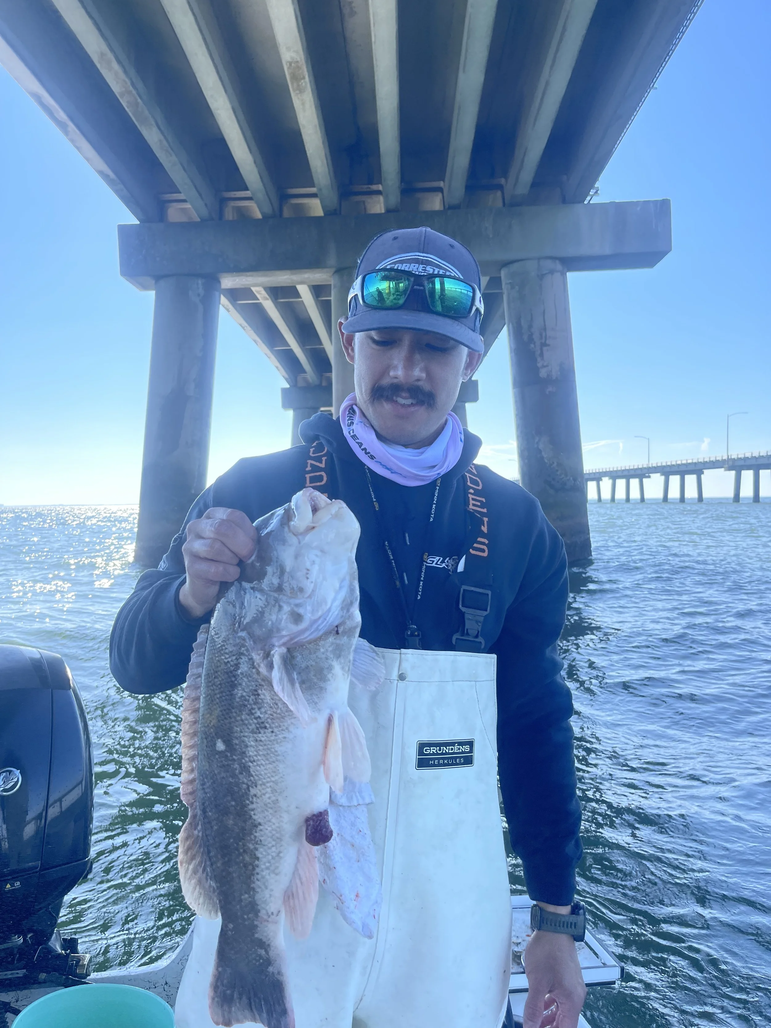 Man holding a large fish under a pier over the water, with a bridge in the background.