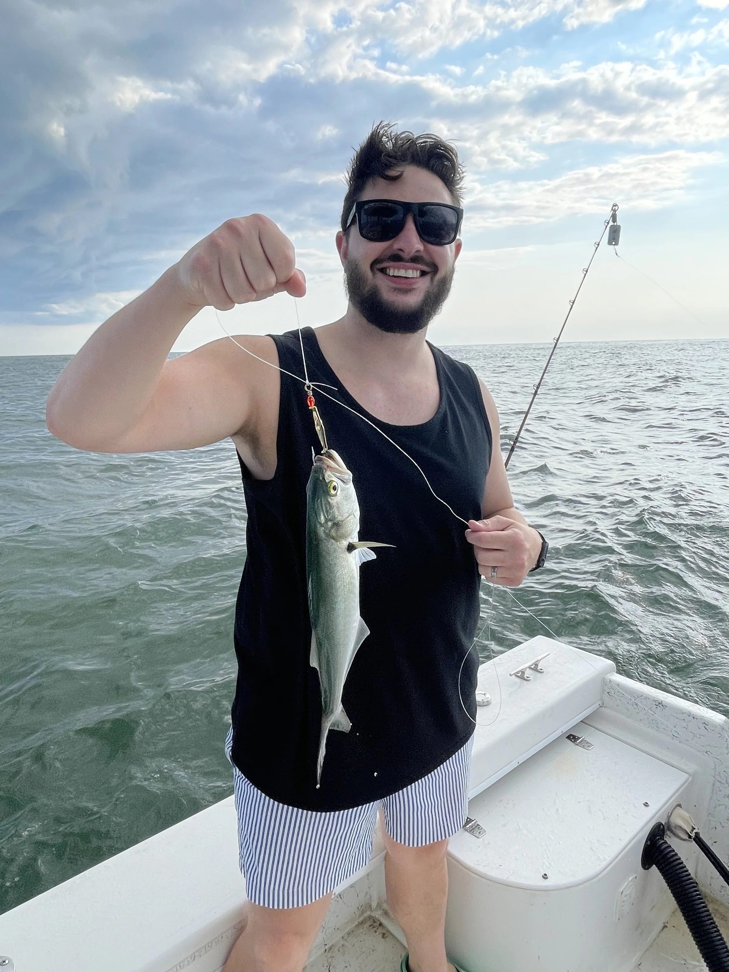 Man holding a fish he caught while fishing on a boat in the ocean, smiling, wearing sunglasses, black sleeveless shirt, and striped shorts, with fishing rod in the background.