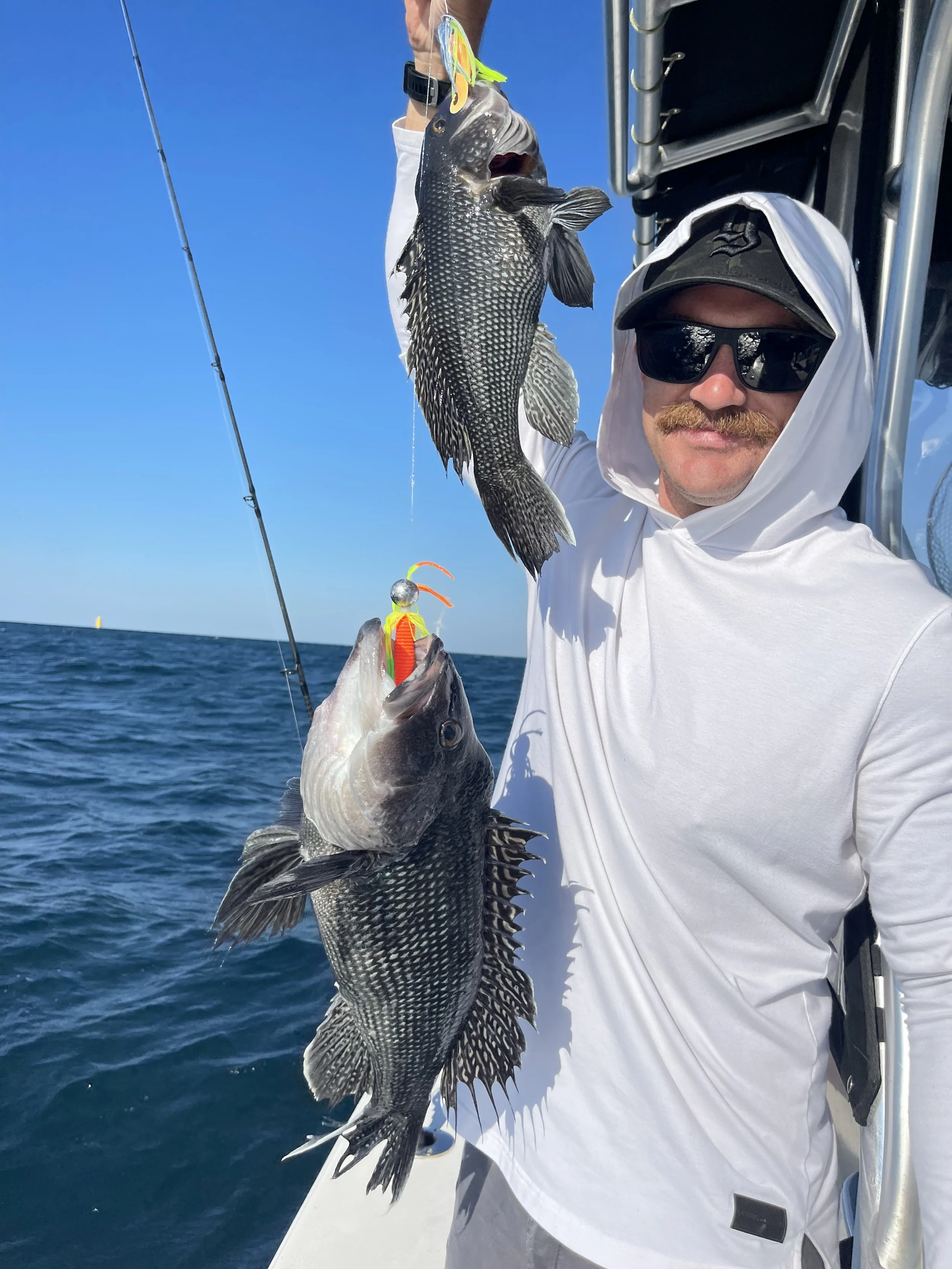 A man with a mustache wearing sunglasses, a white hooded shirt, and a cap, holding two large black and gray fish with fishing lures on their mouths on a boat in the ocean with a clear blue sky in the background.
