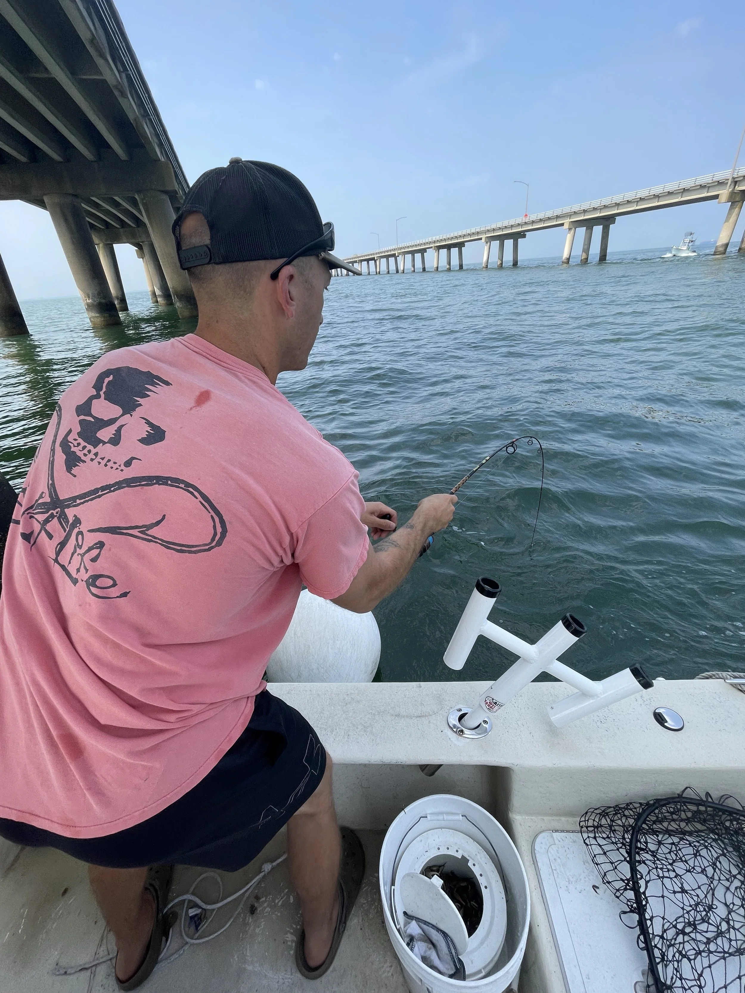 A man wearing a pink T-shirt and black shorts fishing from a boat under a bridge.
