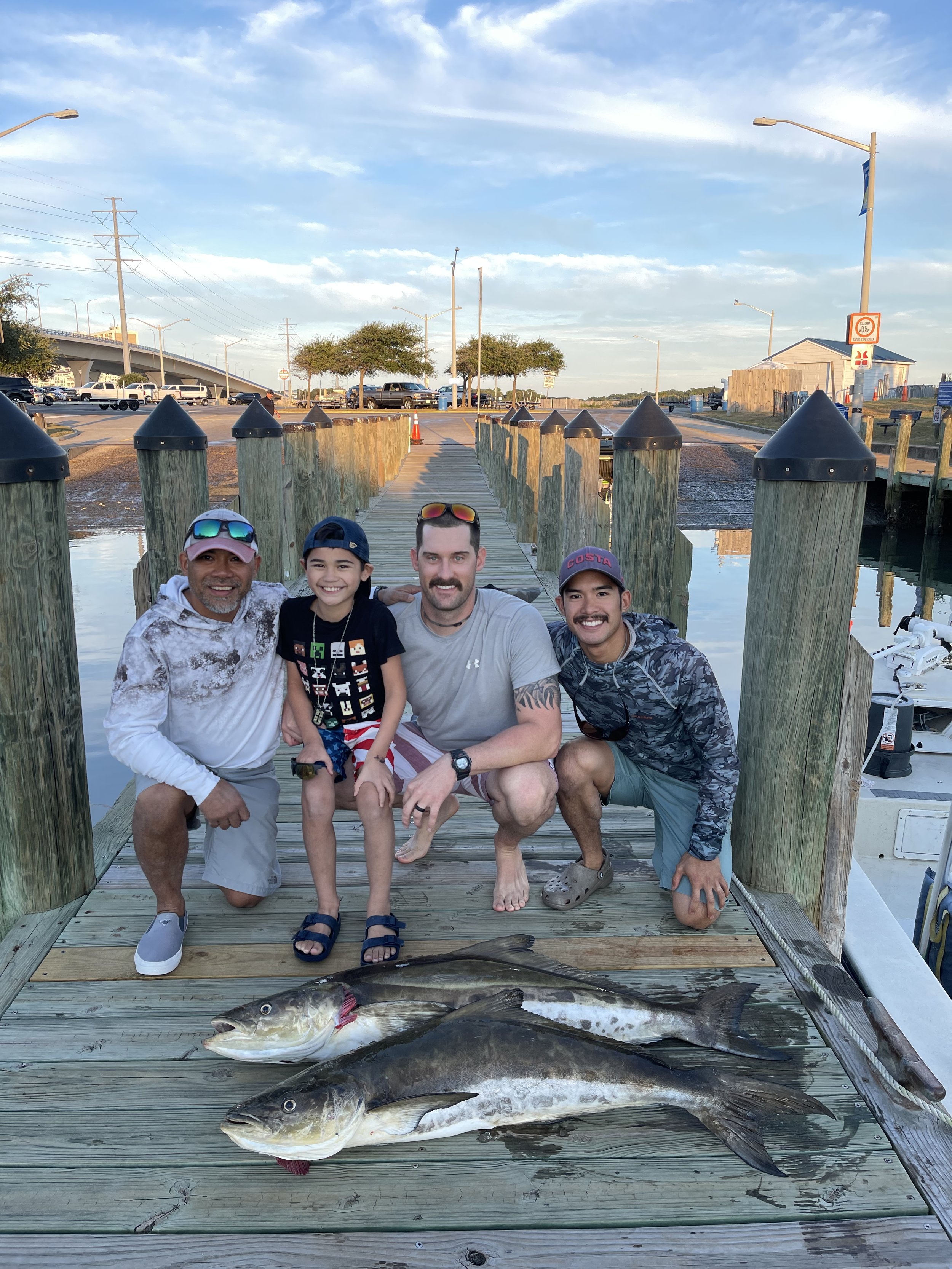 Four people, including a young boy, posing on a dock with two large fish they caught, with boats and water in the background.