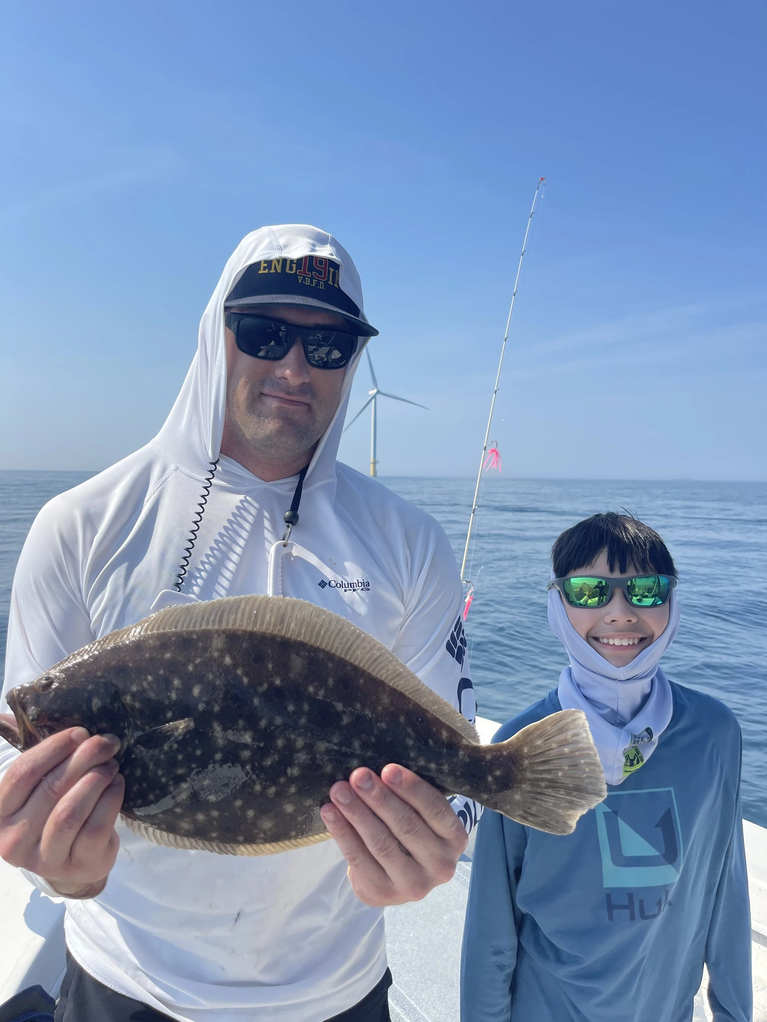 A man and a boy on a boat holding a fish, with a wind turbine and fishing pole in the background.