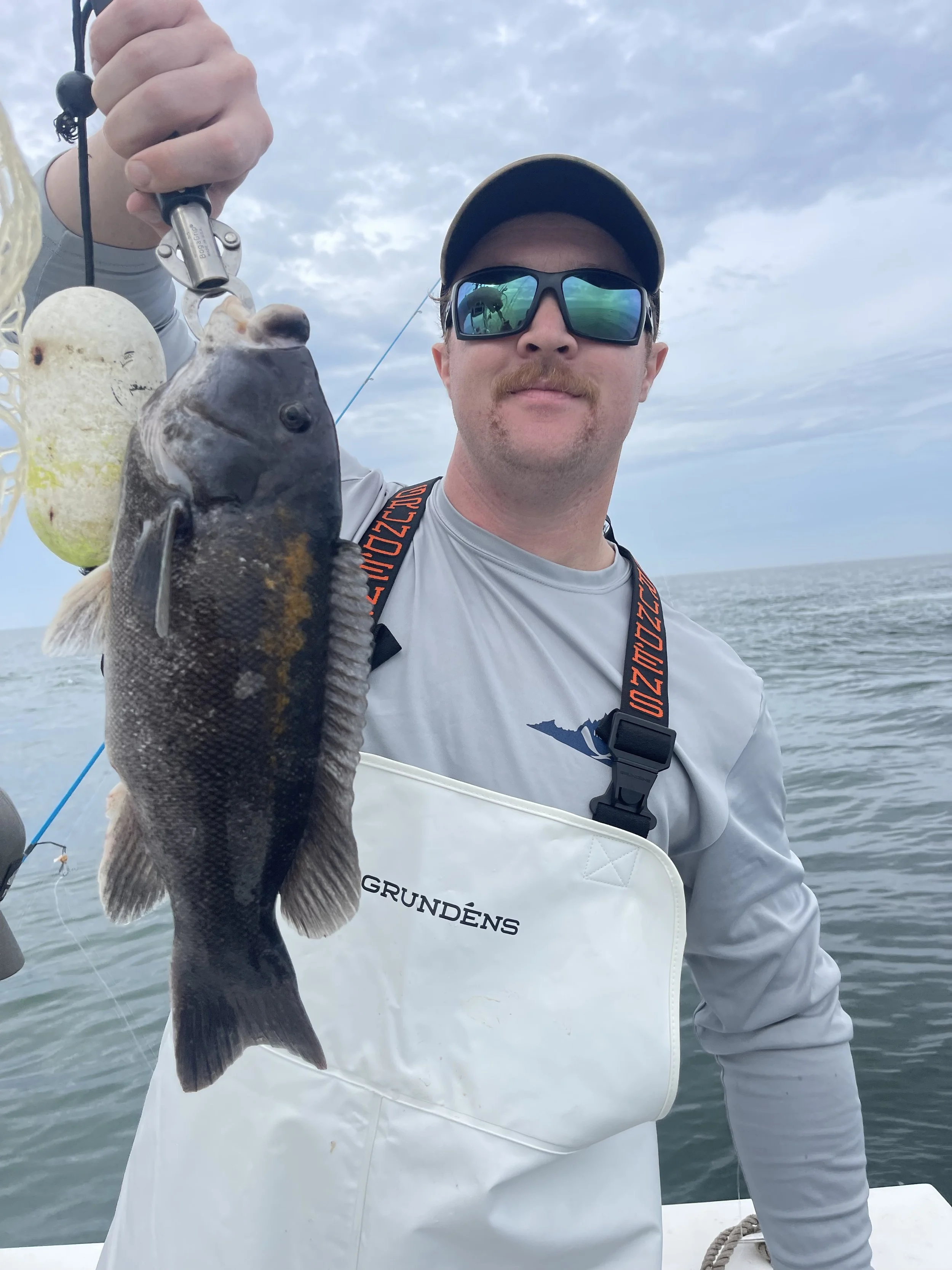 A man with sunglasses and a cap holding a fish he caught on a fishing line, standing on a boat with an ocean background and cloudy sky.