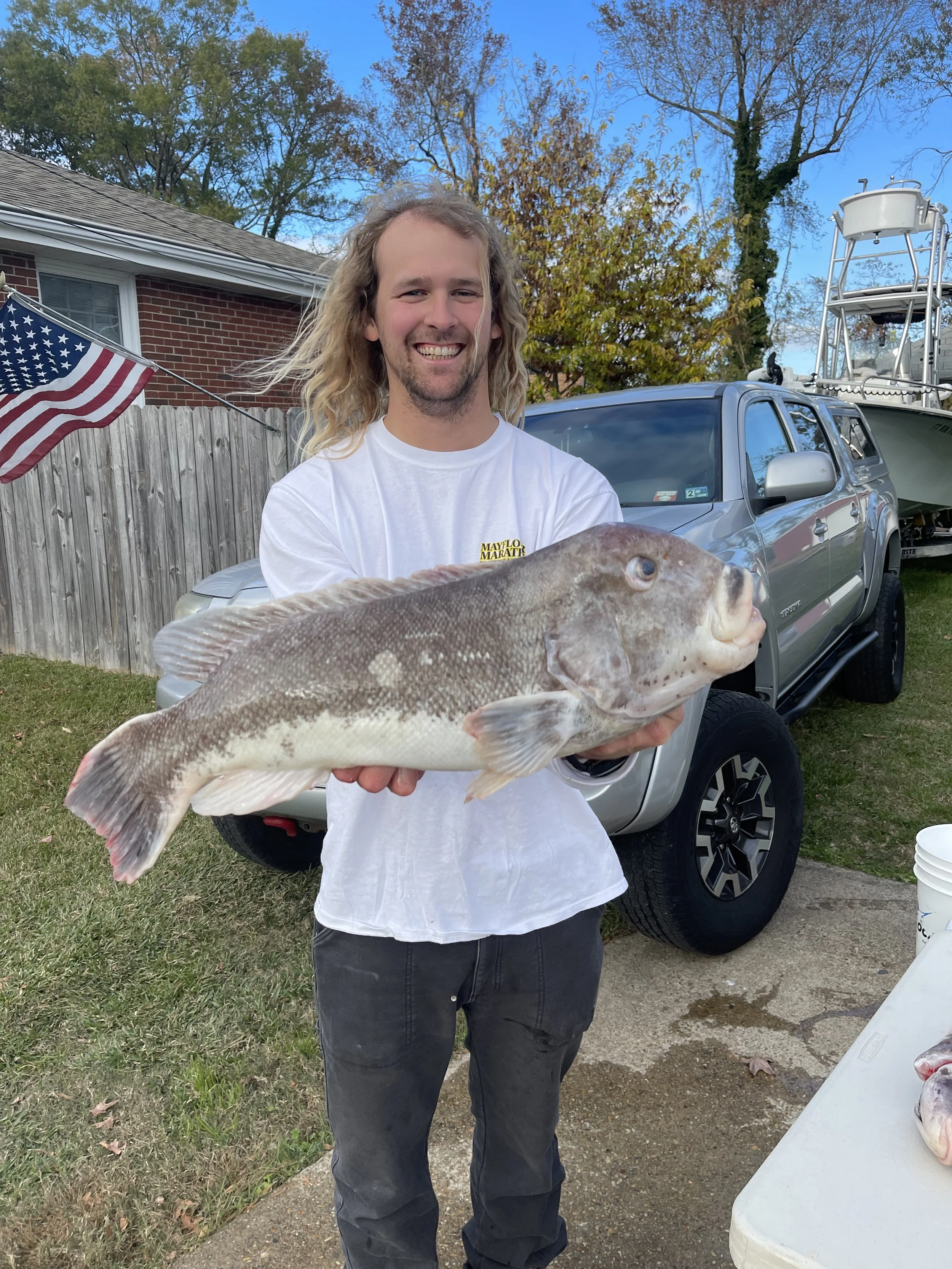 A man with long blonde hair and a beard is smiling while holding a large fish in his hands outdoors. Behind him, there is a parked gray truck, a boat on a trailer, a wooden fence, a house with a brick wall, and a blue sky with some trees.