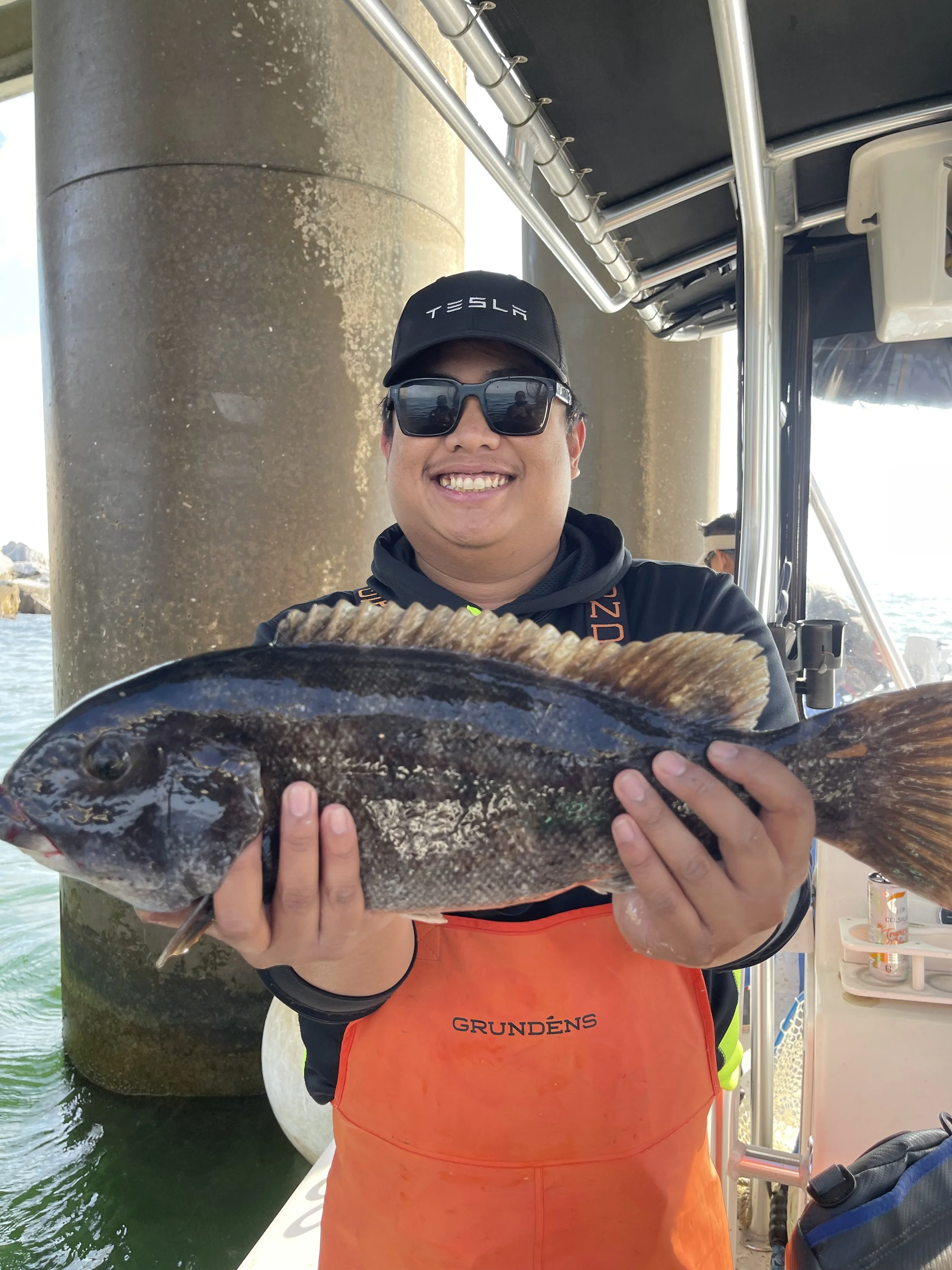 Man wearing sunglasses, a black Tesla cap, and an orange apron is smiling and holding a large fish on a boat.