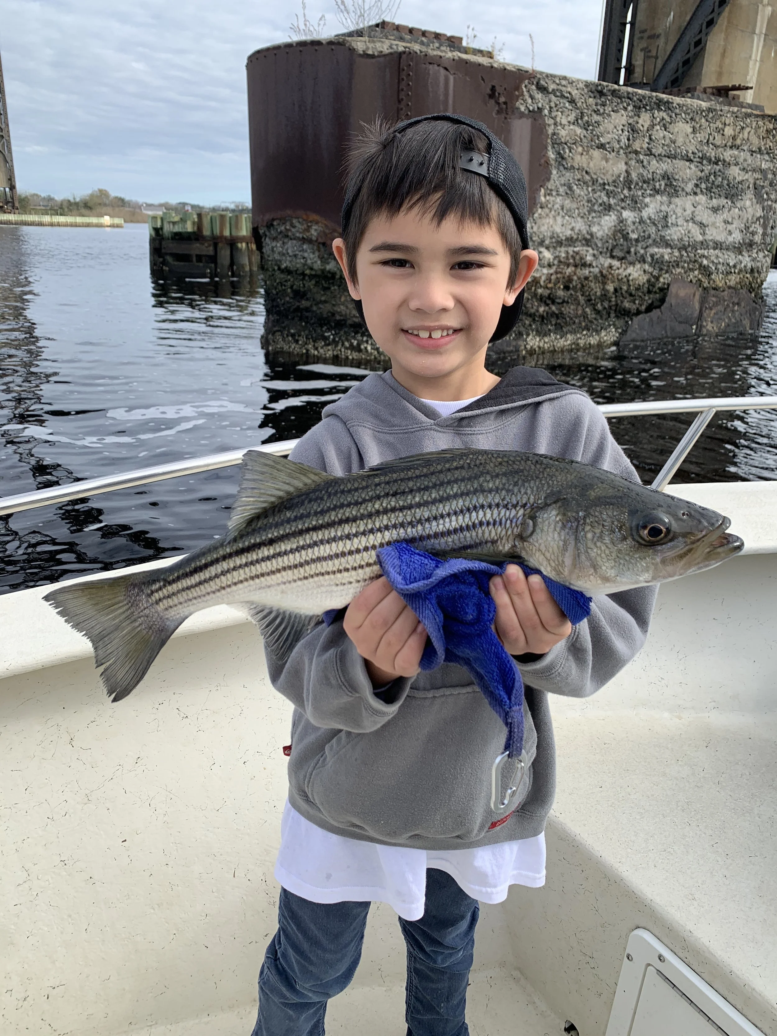 Young boy on a boat holding a large striped bass fish with a blue towel, smiling with a body of water and a large rusted shipwreck in the background.