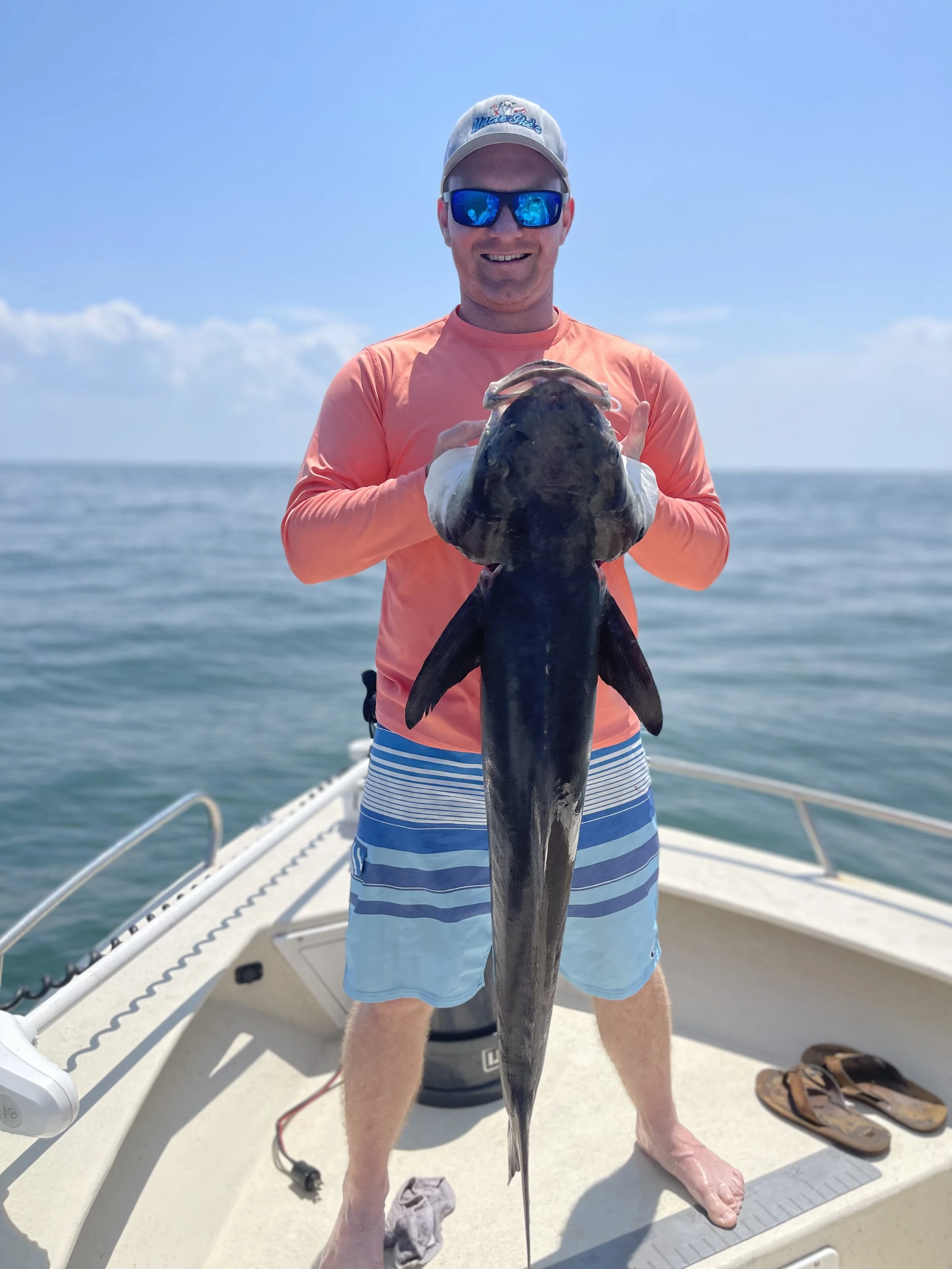 Man on a boat holding a large fish, wearing sunglasses, a hat, an orange long-sleeve shirt, and striped shorts, with the ocean in the background.
