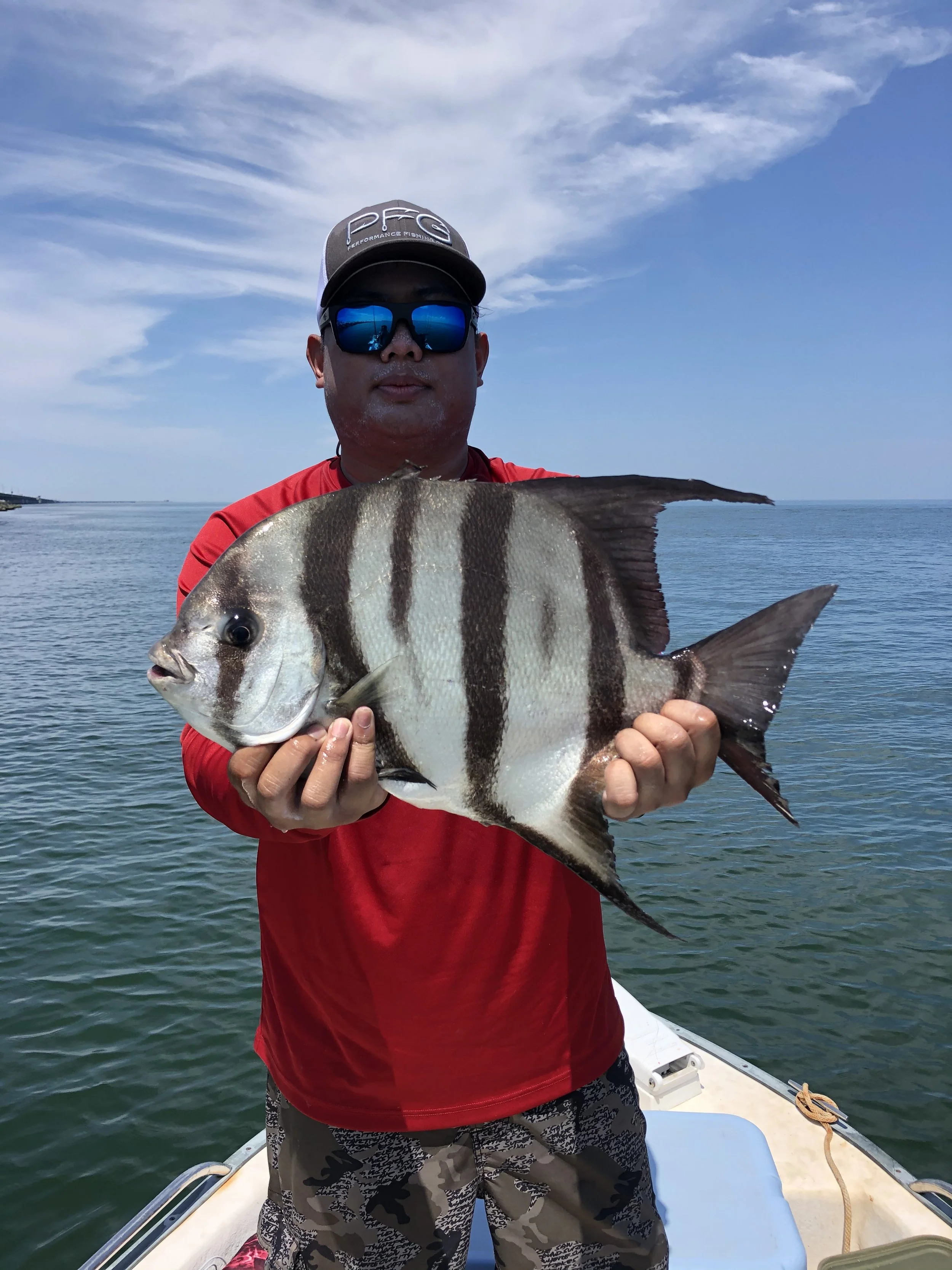 A man wearing a cap, sunglasses, and a red shirt is holding a large striped fish on a boat with water and a cloudy sky in the background.