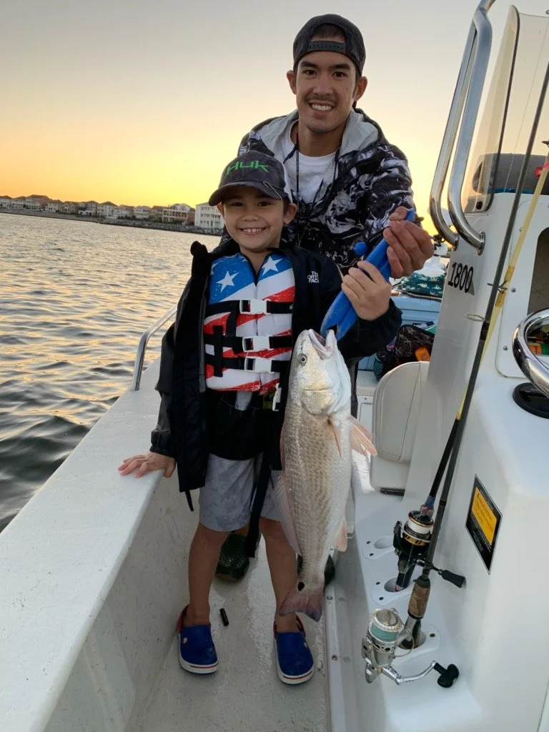 A man and a boy on a boat holding a large fish caught during sunset with water and houses in the background.