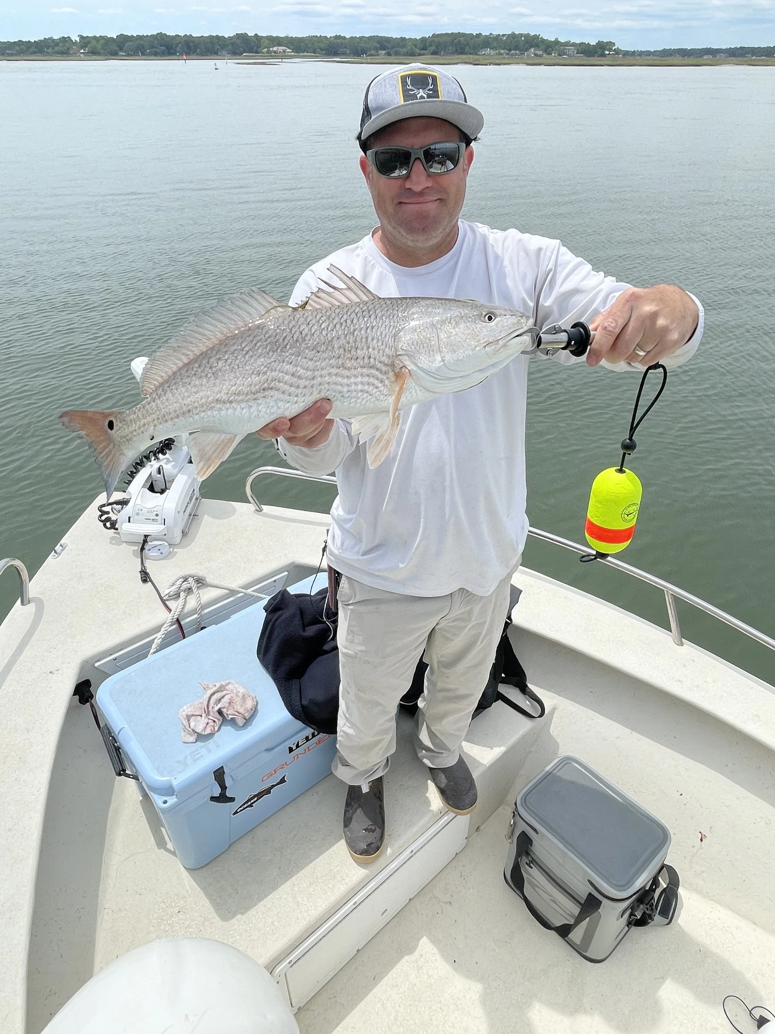 Man on boat holding a large fish with a fishing line and sinker, wearing sunglasses, a cap, and casual clothing, with water and distant shoreline in background.