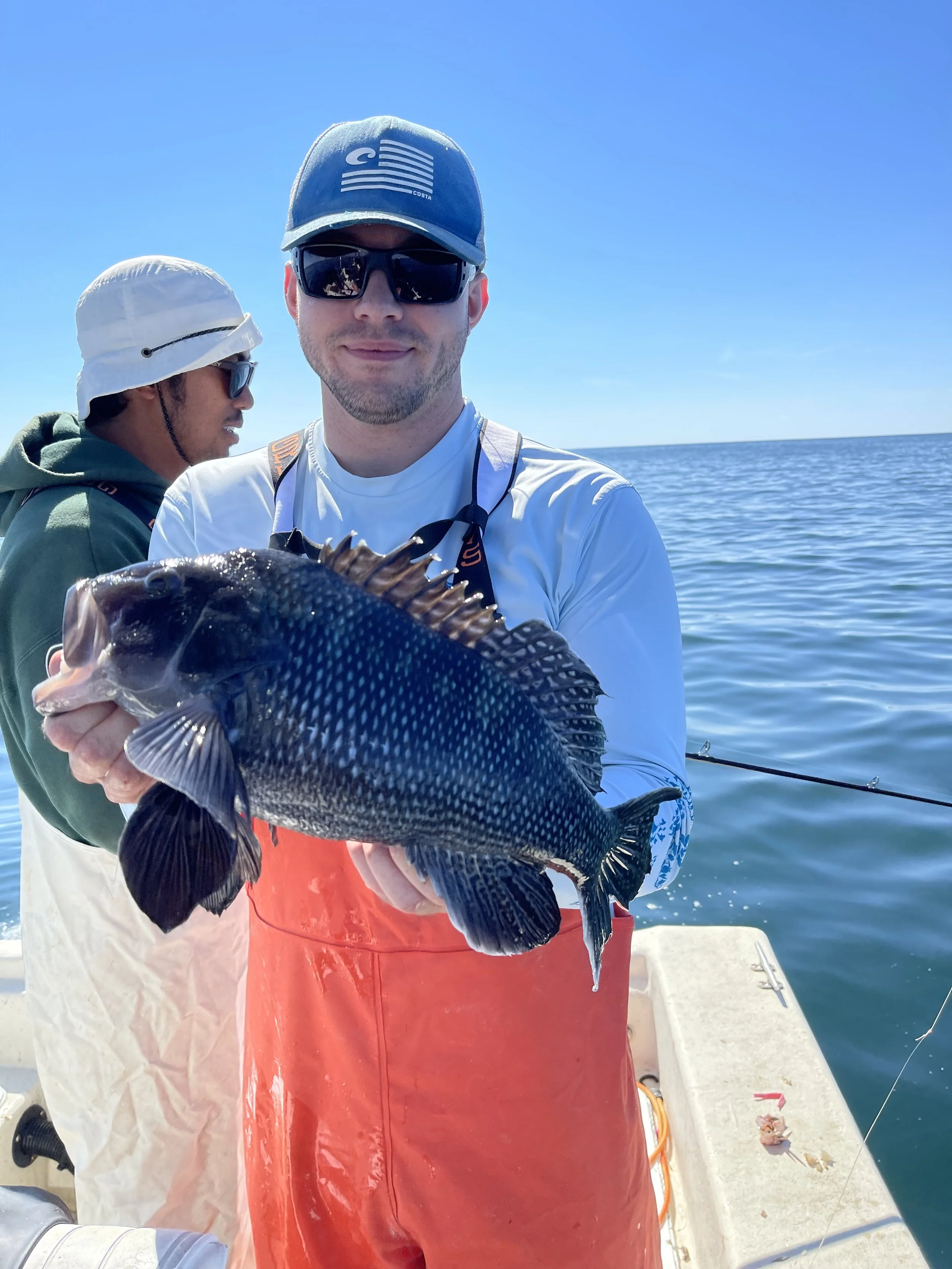 Man in sunglasses and a cap holding a large fish on a boat with ocean in the background, another man in a hat appears in the background.