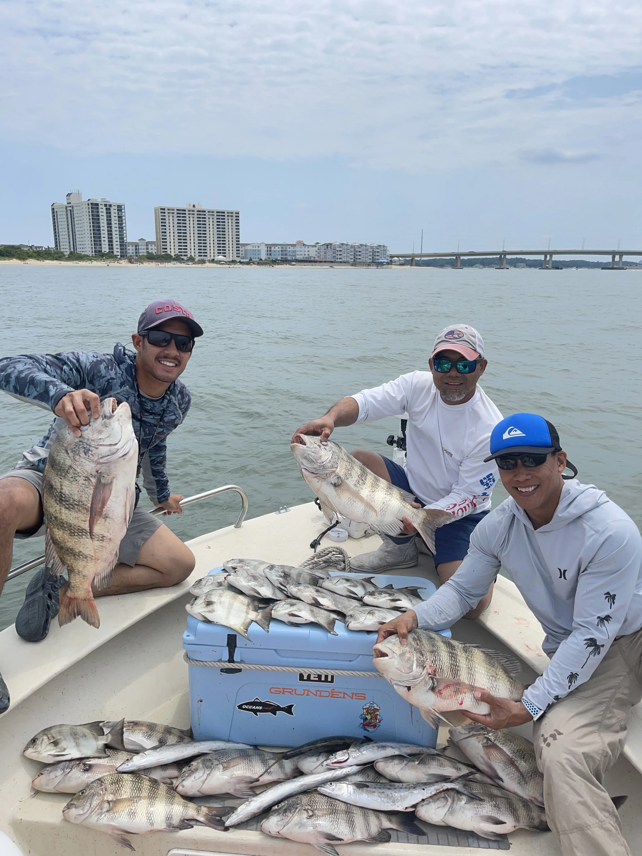 Three men on a boat displaying their fishing catch, including several fish laid out on a cooler and held up by the men, with a cityscape and bridge in the background.