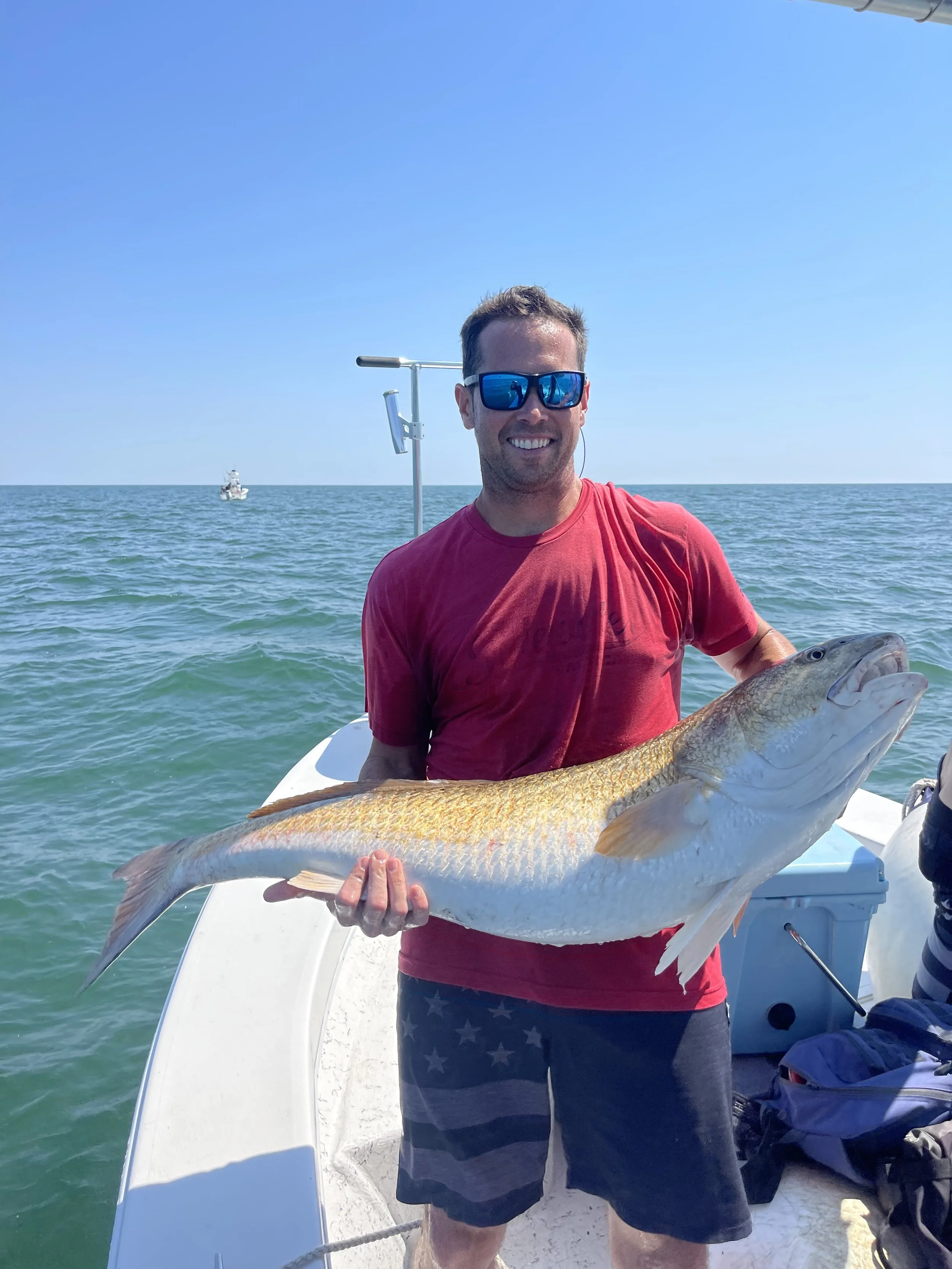 A happy man wearing sunglasses and a red t-shirt holding a large fish on a boat in the ocean with a clear blue sky.