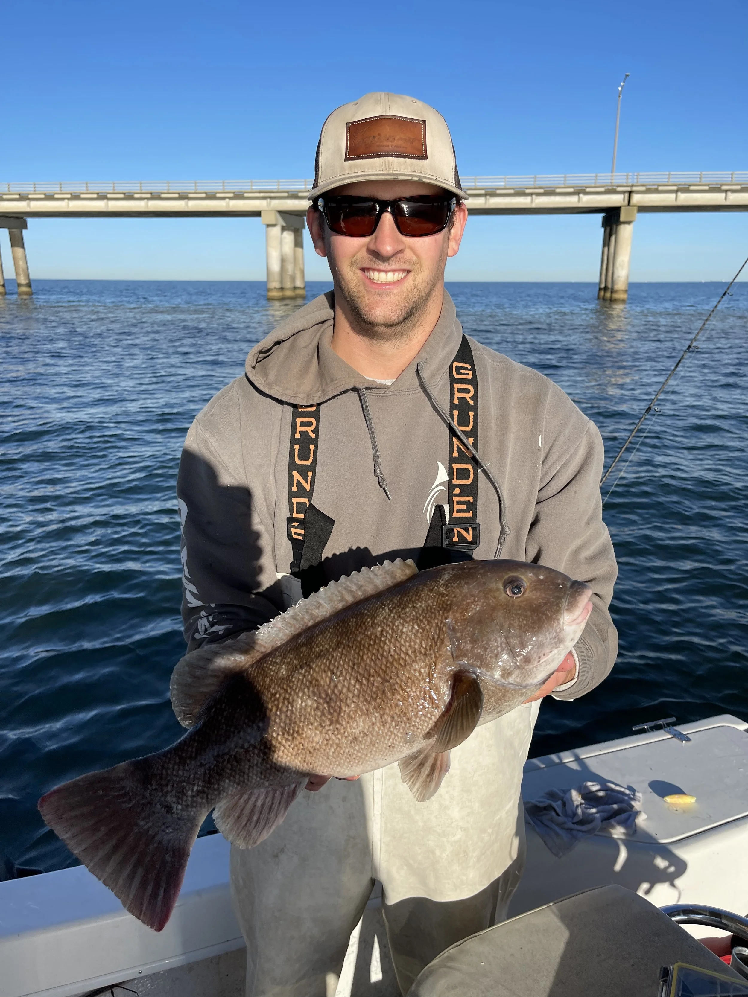 A man wearing sunglasses, a beige cap, and a gray hoodie holding a large fish on a boat, with a bridge and blue water in the background.
