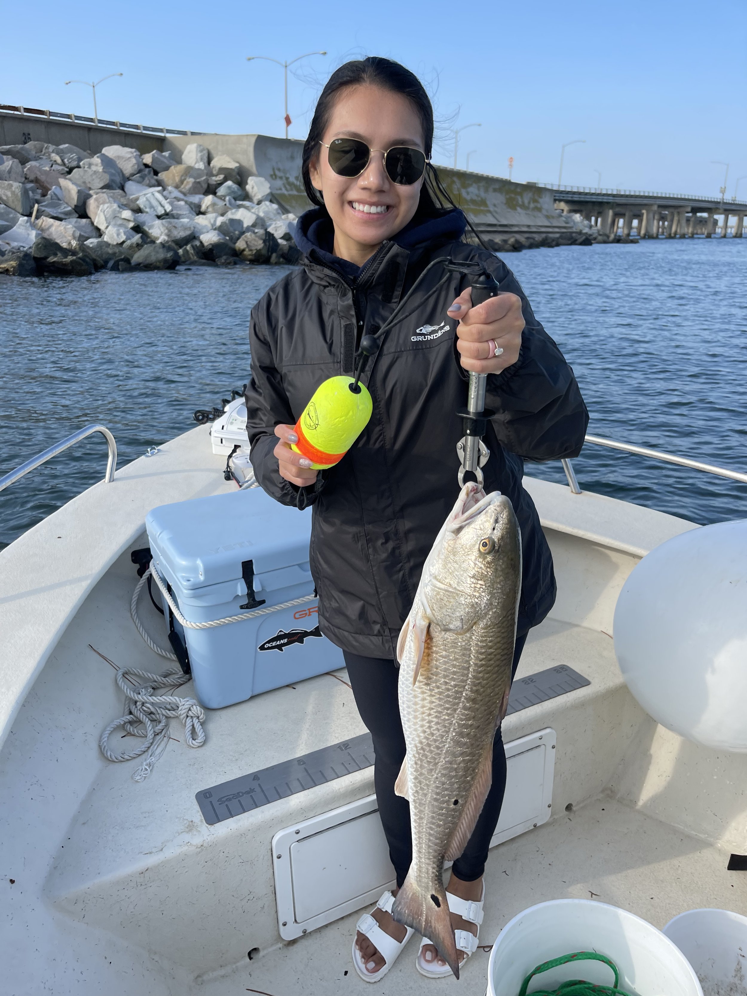 A woman smiling on a boat holding a large fish she caught, wearing sunglasses, a black jacket, and sandals, with a rocky shoreline and bridge in the background.