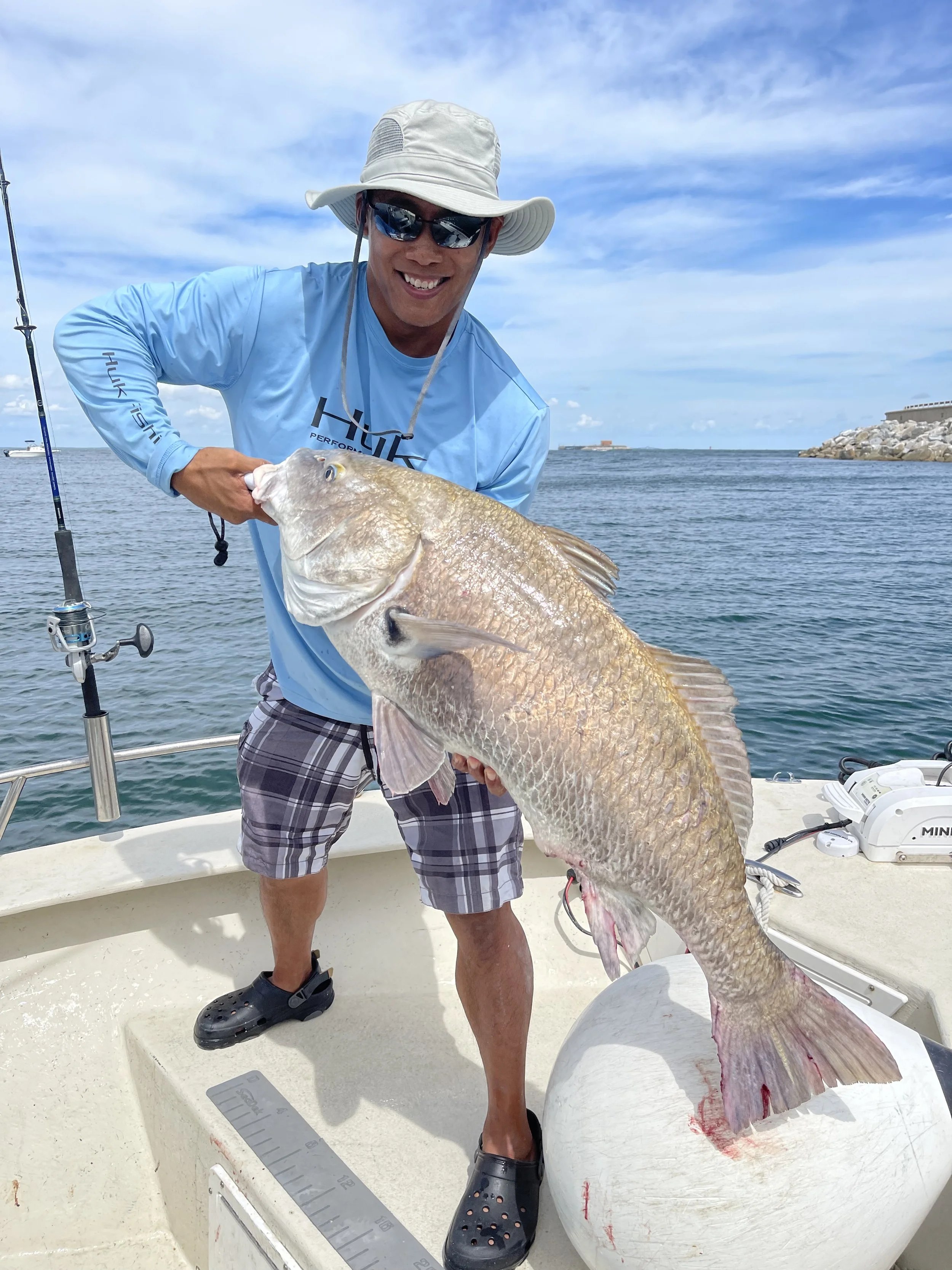 Man on a boat holding a large fish he caught, with a fishing rod and water in the background.