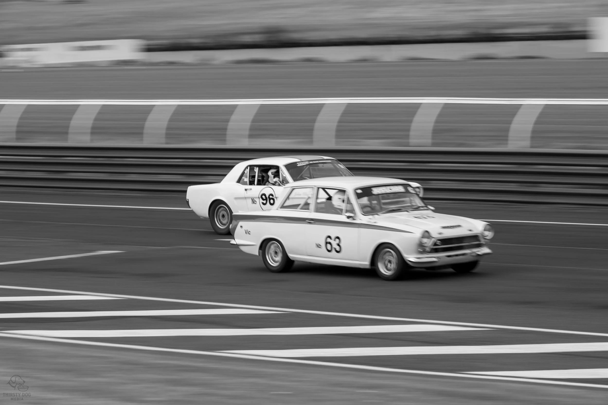 Two vintage race cars competing on a race track, with a brick or stone wall and tire barriers in the background, black and white photograph.
