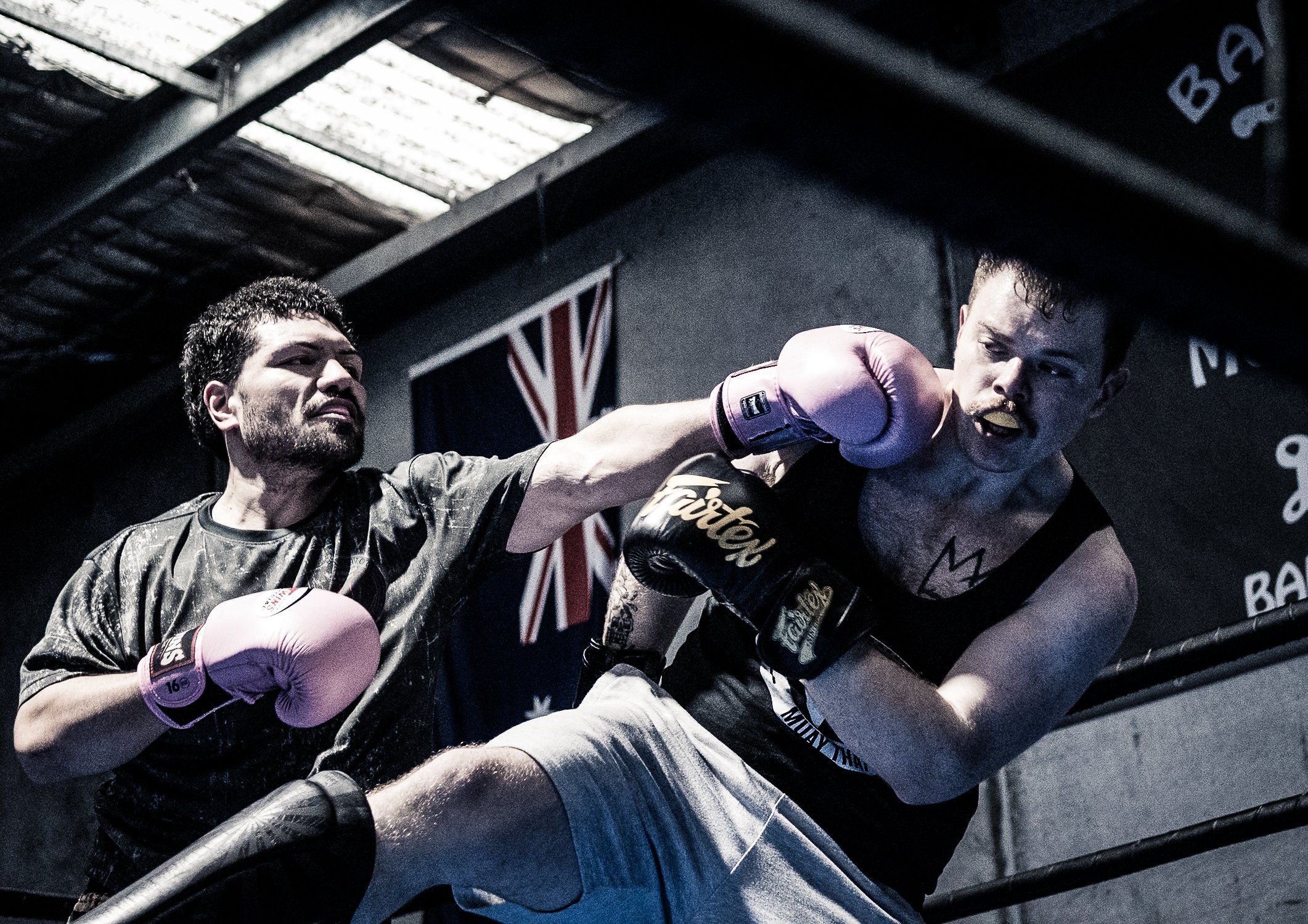Two men engaged in a boxing match inside a gym, with one delivering a punch to the other's face, while both wear boxing gloves and athletic clothing.
