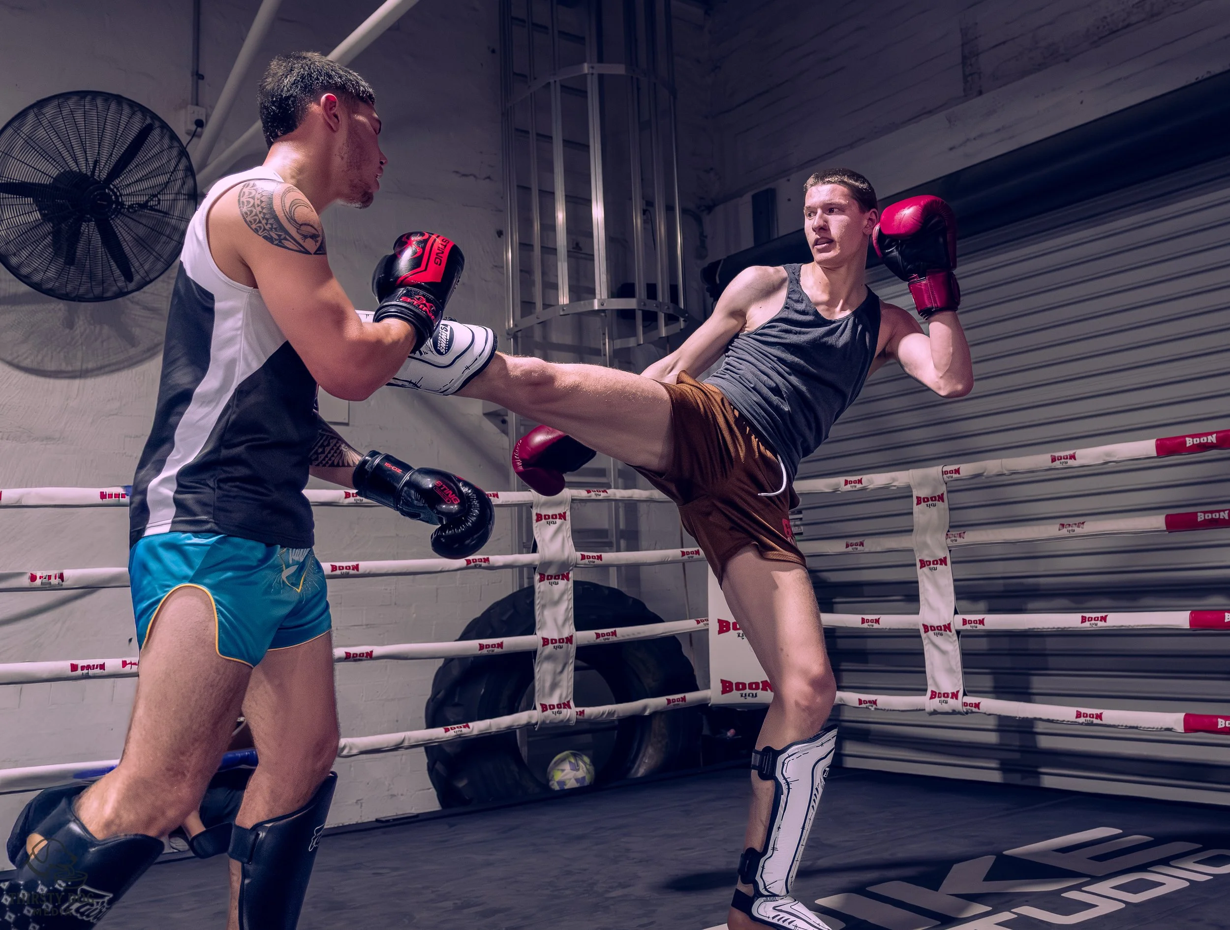 Two men sparring in a boxing ring, one delivering a kick while the other defends with gloves, inside a gym.